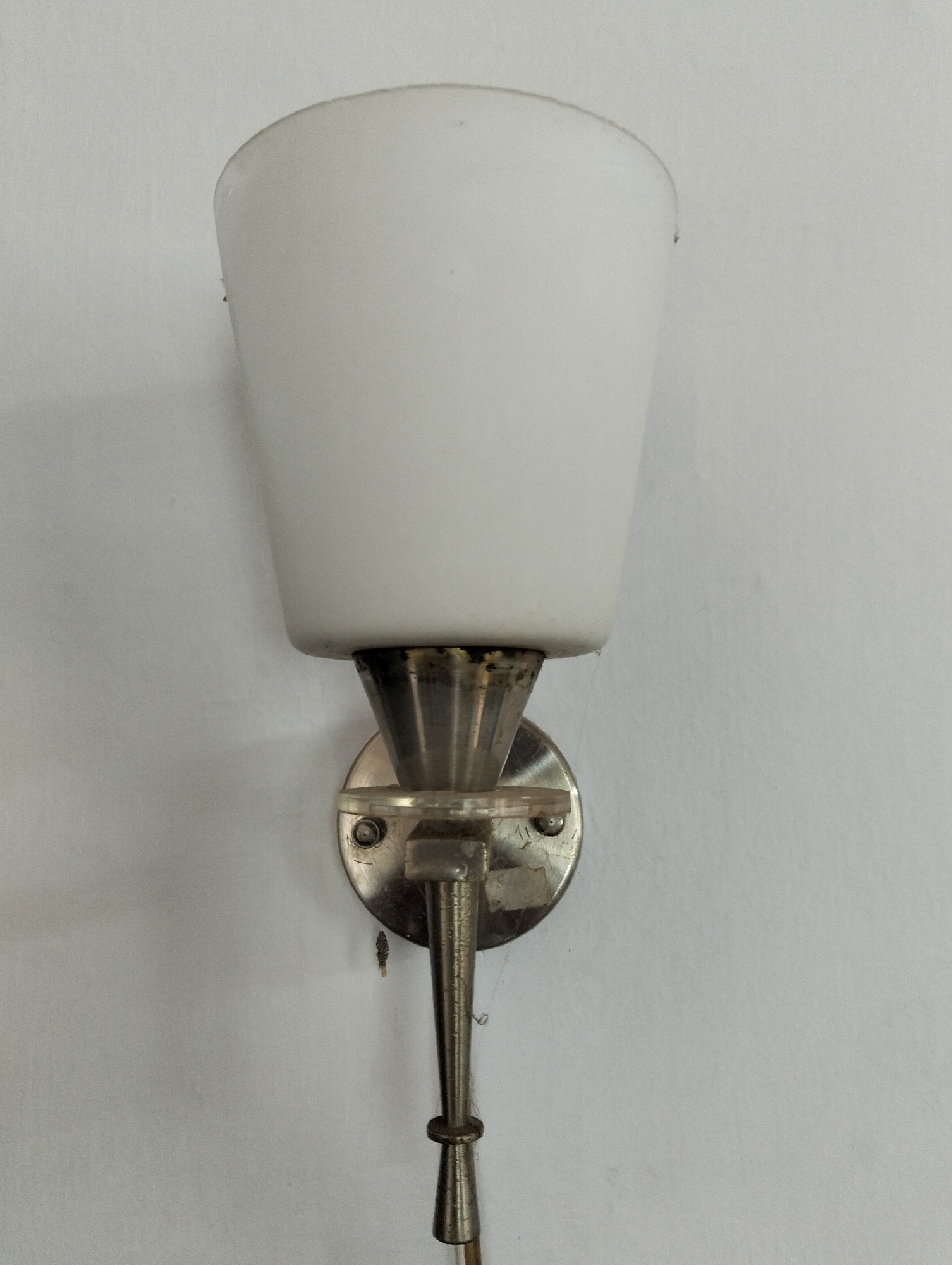A white, conical lampshade is mounted on a brushed metal fixture attached to a white wall. The fixture has a circular base with visible screws, and a slender, decorative arm that extends downwards. The overall appearance suggests a vintage or retro style. The image was taken in Julicotmeurak, Indonesia. There are no people or discernible actions present in the scene.