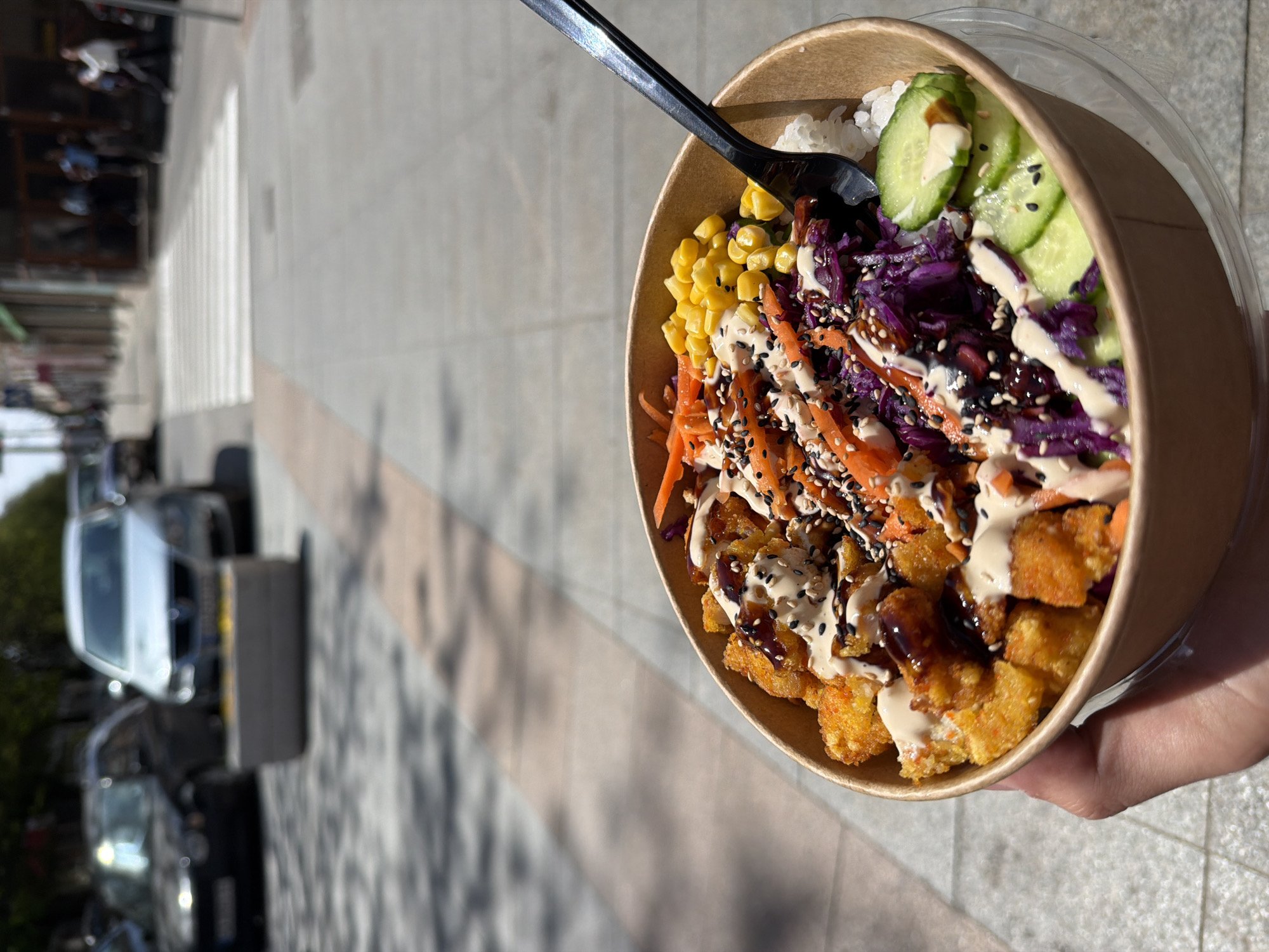 A medium-shot, portrait-oriented, street-level photograph depicts a bowl of food in the foreground, held in someone's hand, with an urban street scene in the background. The bowl contains rice, cucumber slices, shredded carrots, red cabbage, corn, and fried pieces, all drizzled with two different sauces and sprinkled with sesame seeds. A black plastic spoon is stuck into the food. The bowl is made of brown paper. The background shows a sidewalk made of light brown paving stones, a street with parked cars, and buildings on the far side of the street. The photograph is taken outdoors on a sunny day, with shadows visible on the sidewalk. There is no evidence the scene is in Panagyurishte, Bulgaria.