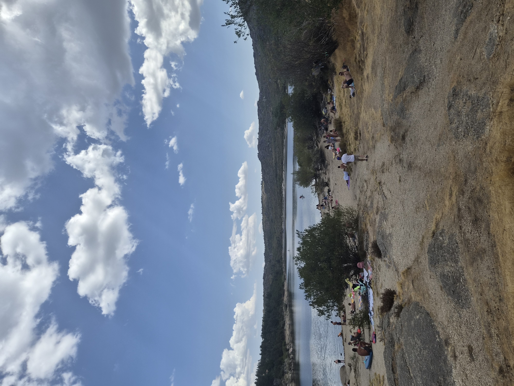 This is a vertical, eye-level, medium shot of a lakeside beach area in Gouveia, Portugal, on a sunny day. The sky is a brilliant blue with scattered white cumulus clouds. The lake is calm and reflects the sky. The beach is sandy with scattered rocks and dry vegetation. A group of people are sunbathing and relaxing on towels and blankets on the beach. Some are lying down, while others are sitting. One person is standing near the water's edge, and several more are in the lake, with at least one on a paddleboard. In the background, a line of trees and rocky hills forms the horizon.