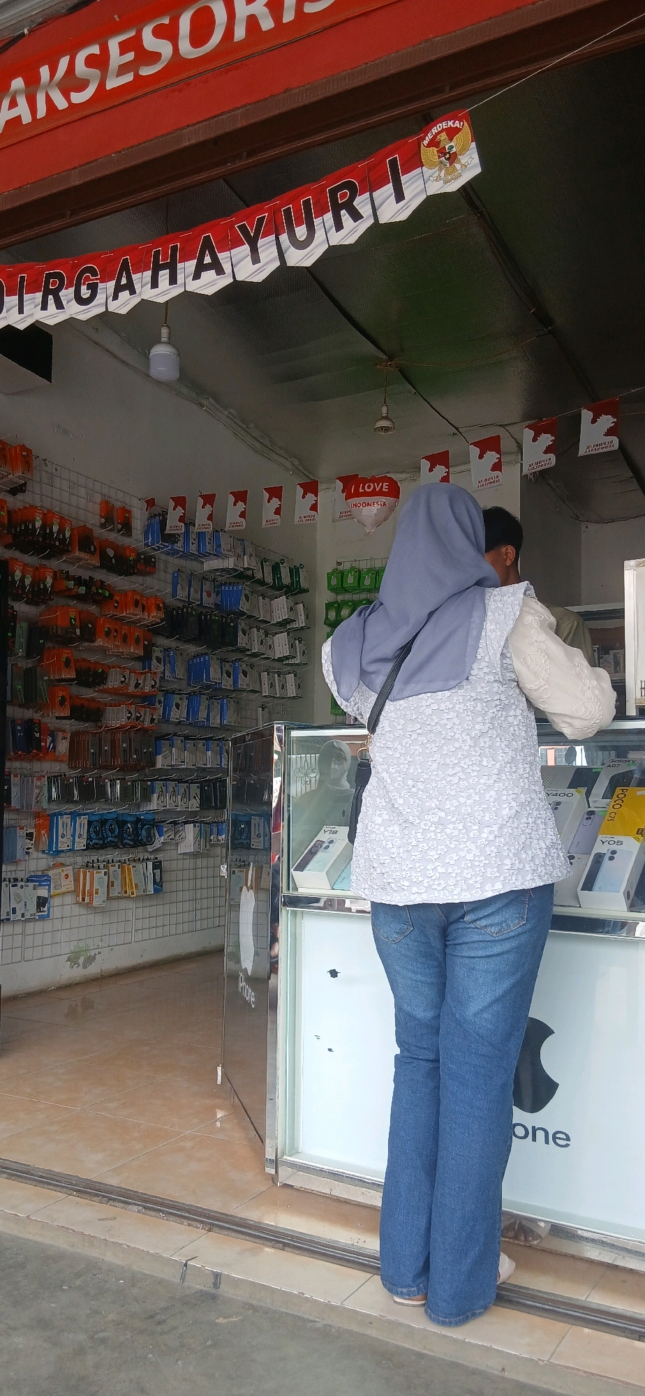 The image shows the exterior of a small shop in the city of Metro, Indonesia, selling mobile phone accessories. In the foreground, a person wearing a blue hijab and blue jeans stands facing away from the camera, looking into the shop. The shop has a display case filled with mobile phones and accessories, and shelves stocked with various phone cases and other items. The shop front is decorated with red and white banners, some featuring the Indonesian flag and the word "MERDEKA," which means "independence." The shop appears to be on a street or sidewalk, with a tiled floor inside and a concrete exterior. The overall impression is that of a bustling local business.