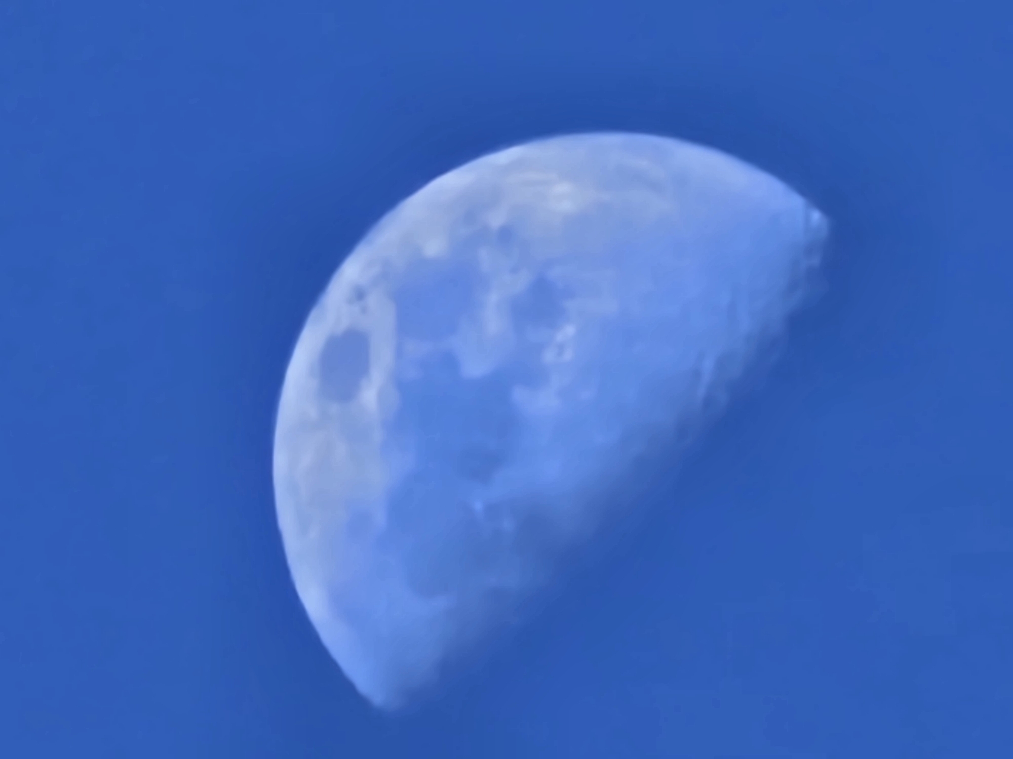 The image displays a celestial body, identified as the moon, in a half-phase (first or third quarter). The illuminated portion of the moon is visible on the right, exhibiting a predominantly cool blue-white coloration with visible dark and light surface features indicative of lunar topography, rendered with soft focus. The moon is positioned against a clear, uniform deep blue sky, devoid of clouds or other atmospheric phenomena. No other objects, individuals, or environmental details from the terrestrial landscape are discernible. The scene depicts the moon as observed from Esch-sur-Alzette, Luxembourg.