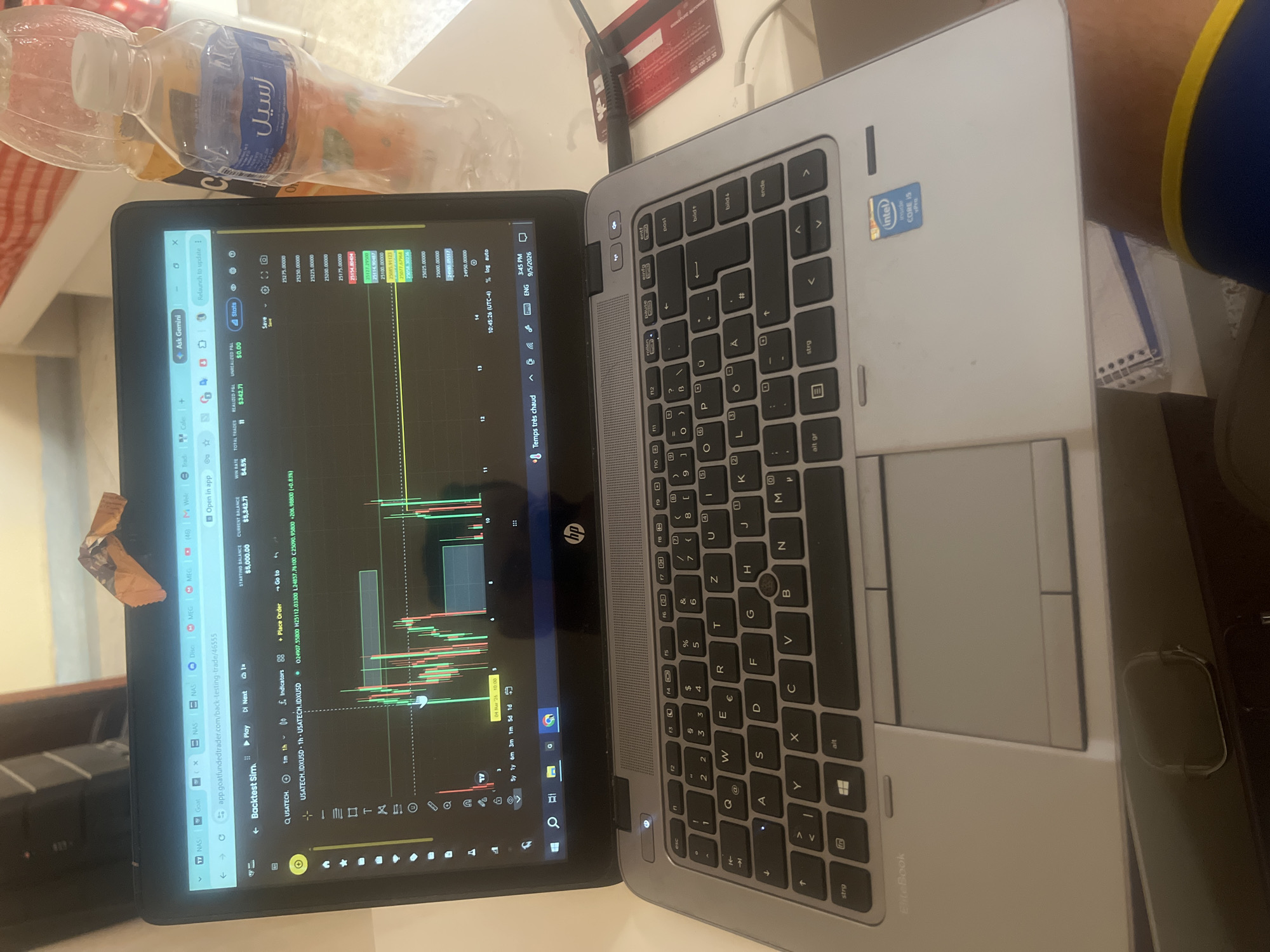 An overhead, close-up shot captures a black HP EliteBook laptop open on a table, displaying a trading platform with a candlestick chart. To the right of the laptop, a partially obscured plastic water bottle with Arabic writing on its label, "أسيل," and an orange-colored drink bottle are visible. A red credit card is placed on the table, connected to the laptop via a cable. The laptop is in the city of Ouaouizert, Morocco, suggested by the Arabic text on the water bottle and the overall context of the image. The scene is likely indoors, with a blurred background that hints at a room with striped bedding. The lighting suggests it is daytime.