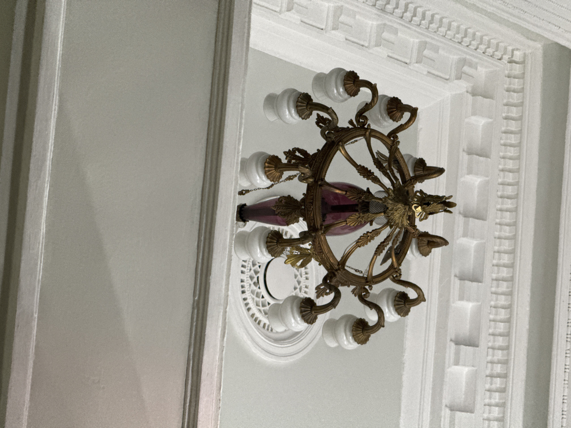 A close-up interior shot depicts a chandelier affixed to a white ceiling. The chandelier, composed of a central red glass section and golden, leaf-like arms supporting white, bulbous lights, hangs centrally in the frame. The ceiling is adorned with white molding, featuring a repeating geometric pattern. To the left of the ceiling, a portion of a gray wall is visible, bisected by a white door frame. The lighting in the shot is soft, contributing to a clean, bright aesthetic. Location and time are unknown.
