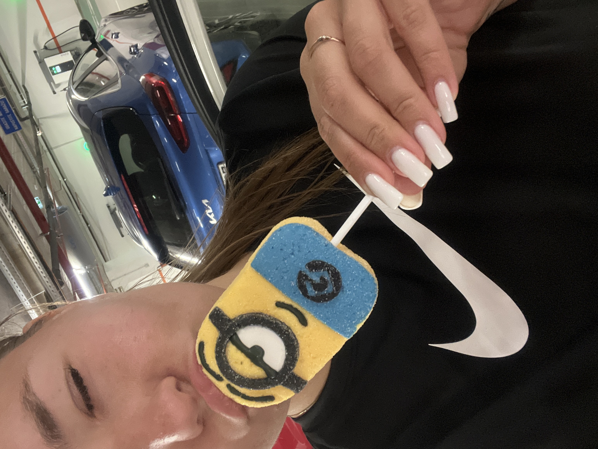 The image shows a person holding a Minion-themed marshmallow lollipop. The person's face is partially visible, with one eye closed and lips slightly parted. Their nails are long and white. They are wearing a black top with a white Nike swoosh logo on the chest. A gold ring is visible on one of their fingers. The background includes a blurred blue car, possibly a Kia, parked near a charging station, and some infrastructure with pipes. The location is likely an underground parking garage. While the specific location in Budva, Montenegro, cannot be confirmed from the image, the style of the charging station and parking garage is common in urban areas.