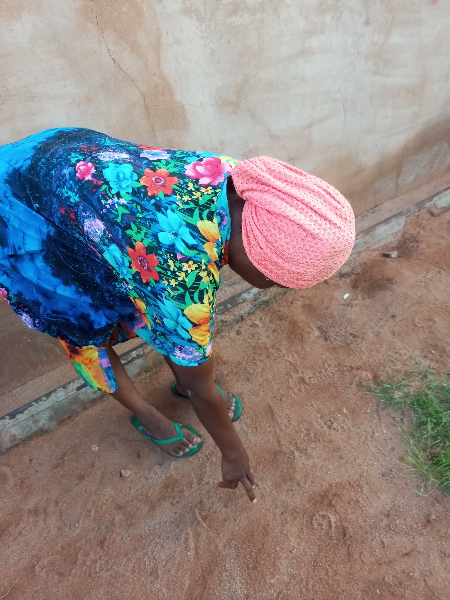A person wearing a colorful floral print dress and a pink headscarf is bending down in an outdoor area. The person is wearing green flip-flops. The ground is sandy and there is a wall in the background. The scene appears to be in Tempure, Nigeria.