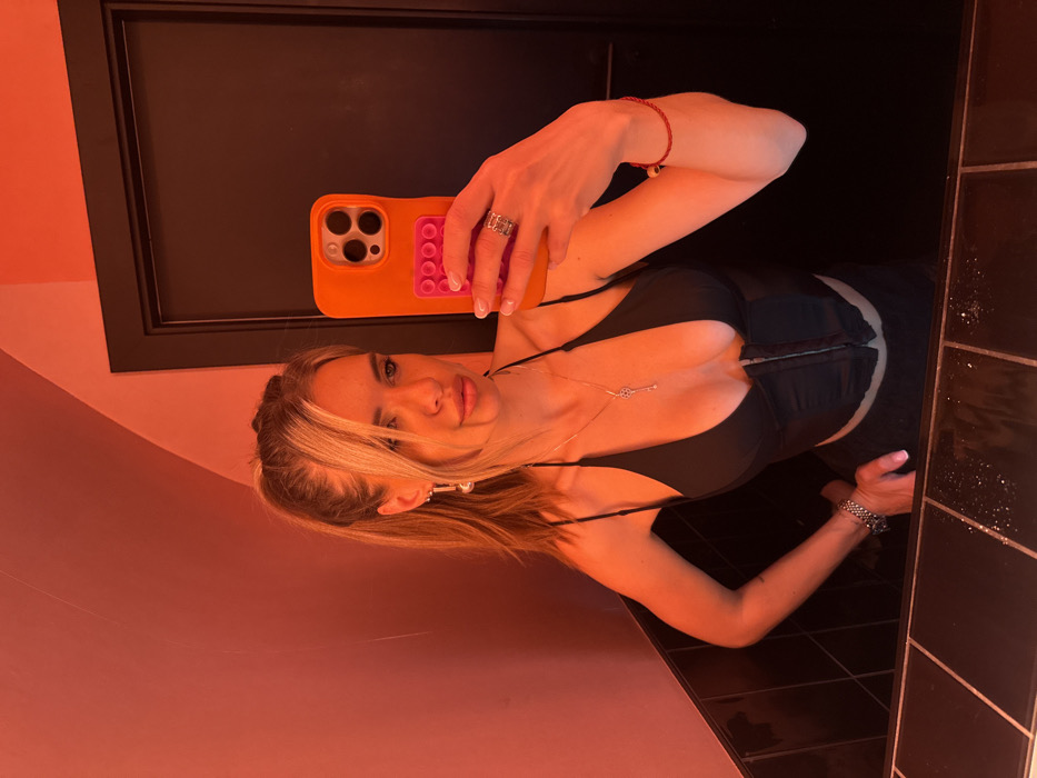 A woman with blonde hair takes a mirror selfie. She wears a black top and a silver necklace. An orange phone case is visible. Her arms and hands feature bracelets and a ring. The mirror reflects a room with pink walls, a black door, and black tiles.