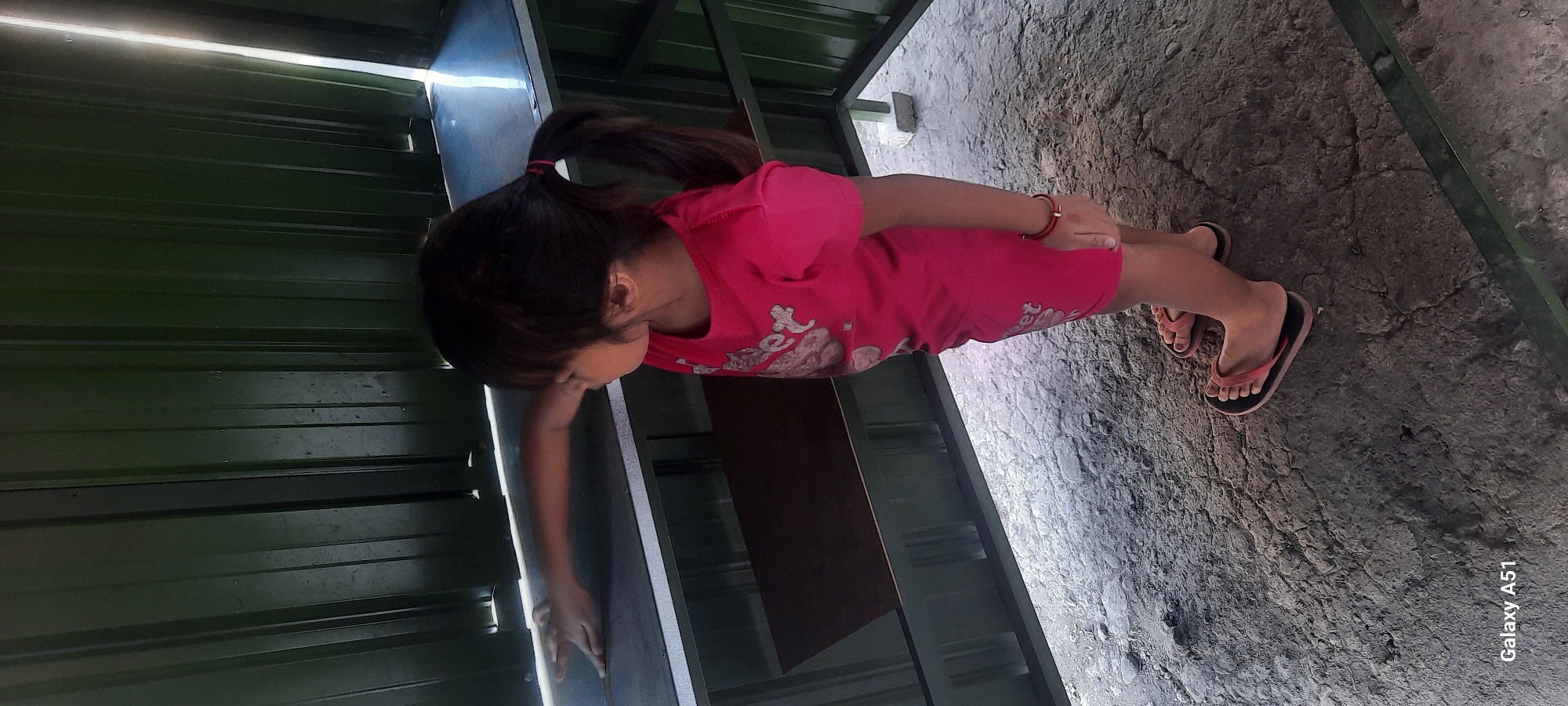 In Selopuro, Indonesia, a young girl with dark hair pulled into a ponytail, wearing a bright pink t-shirt and pink flip-flops, stands inside a metal structure. The structure appears to be a shelter or storage unit with corrugated green metal walls. The girl's left arm is extended, reaching towards a shelf within the structure. The ground is a rough, textured surface that appears to be a mix of dirt and gravel. Sunlight is visible, casting shadows and highlighting parts of the interior. The overall scene suggests a casual, outdoor setting.