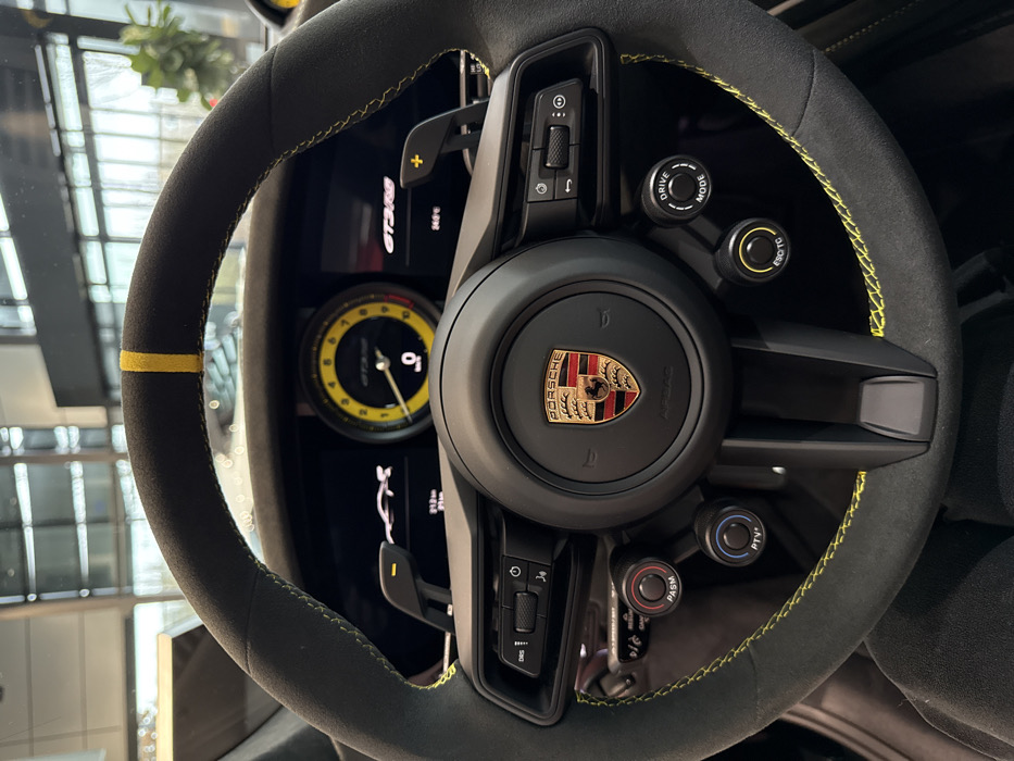 The image provides a detailed view of the cockpit of a Porsche 911 GT3 RS, observed from the driver's perspective, likely situated within a car dealership showroom in Regensburg, Germany.
The primary subject is the black Alcantara-wrapped steering wheel, distinguished by bright yellow contrast stitching along its circumference and a yellow marker at the 12 o'clock position. The center of the steering wheel features the multicolored Porsche crest. Integrated controls include left-side buttons for "DRS" and volume, along with two rotary dials labeled "PASM" and "PTV+". The right side of the steering wheel contains control buttons, and two additional rotary dials labeled "DRIVE MODE" and "ESC/TC". Black paddle shifters are visible behind the steering wheel, positioned for gear changes.
Through the steering wheel, the instrument cluster is visible. A prominent central analog tachometer features a yellow face with black markings, displaying "GT3RS" at its base and indicating "0" RPM. Flanking the tachometer are digital displays. The left screen shows a silhouette of the Porsche GT3 RS and an odometer reading of "312.9 km". The right screen reiterates "GT3RS" and displays an ambient temperature of "24.0 C".
A person's dark-clad legs are visible at the bottom of the frame, indicating an individual is seated in the driver's seat. The background, blurred through the windshield, reveals a modern, well-lit interior space with large glass windows and a distant green plant, consistent with the environment of an automotive showroom.