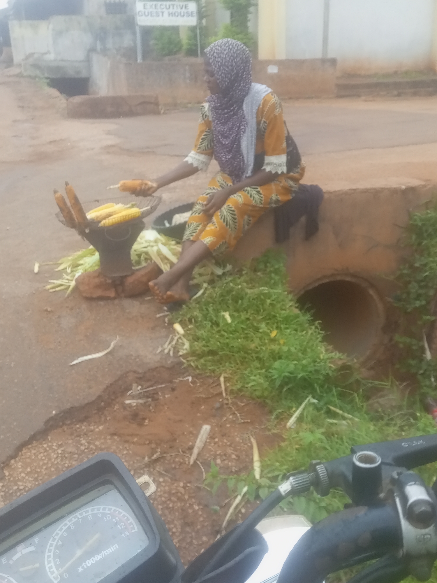 A woman in a hijab and a patterned dress sits on a concrete curb next to a dirt road in an outdoor, possibly urban, setting. She is in the process of roasting corn. A basket of unroasted corn and husks is in front of her, and she holds a roasted ear of corn in her left hand. A black object, possibly a motorcycle's instrument panel, is visible in the foreground at the bottom of the image. Behind the woman is a large concrete culvert with vegetation growing around it. A sign with the words "EXECUTIVE GUEST HOUSE" is visible in the background.