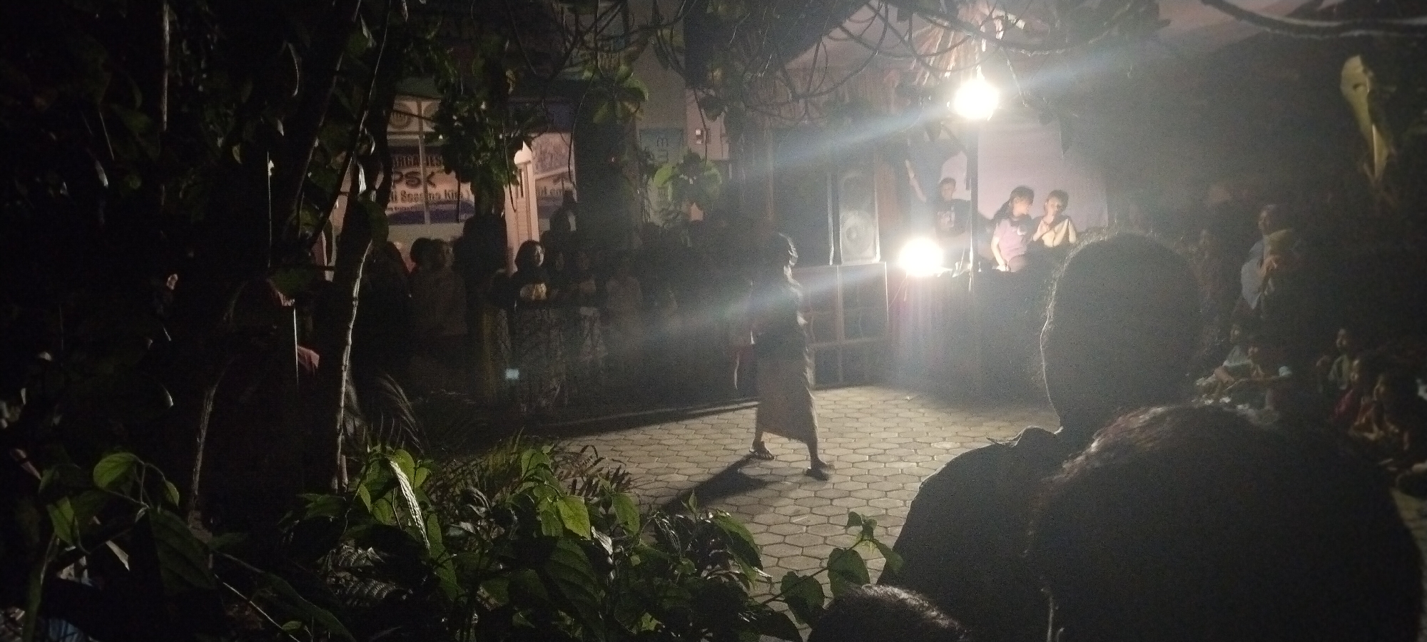 The image depicts an outdoor nighttime gathering in Keting, Indonesia, likely a community event or performance. In the foreground, the silhouette of a person's head and shoulders is visible, suggesting they are an observer. The middle ground features a paved area with a hexagonal pattern, where a lone figure in a light-colored garment is walking or dancing, illuminated by bright stage lights. Behind this performer, large speakers are stacked, indicating a sound system is in use. A group of people are gathered in the background, some seated and others standing, watching the performance. The environment is dimly lit, with natural foliage and branches visible in the foreground and framing the scene. The overall atmosphere appears to be lively, with the bright lights creating a focal point on the performers and the audience engaged in observation.