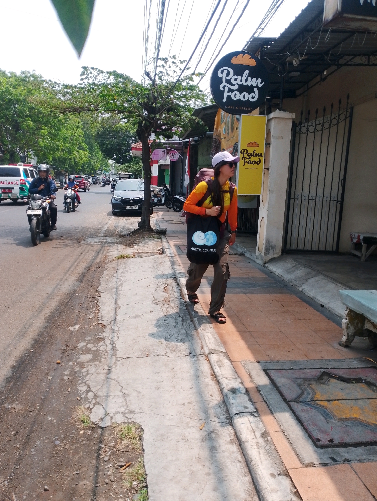 A person walks along a sidewalk in Probolinggo, Indonesia, passing a business with a sign that reads "Palm Food Cake & Bakery." The person is wearing a white cap, sunglasses, a bright orange and yellow long-sleeved shirt, brown cargo pants, and black sandals. They have a large backpack on and are carrying a black tote bag with the "Arctic Council" logo. The sidewalk is paved with tiles and concrete, and there is a drainage grate with a star pattern visible. The street is busy with traffic, including motorcycles and cars. An ambulance is visible on the left side of the street. The area is lined with trees and overhead wires. The lighting suggests it is daytime, with shadows cast on the sidewalk.