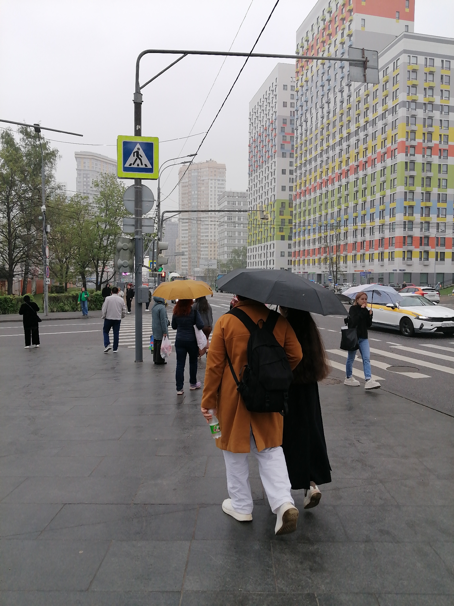 The image captures a street scene in Moscow, Russia, on a cloudy day, as evidenced by the overcast sky and umbrellas. Several pedestrians are crossing the street at a crosswalk, indicated by the blue and yellow pedestrian crossing sign. Some people are holding umbrellas, including a yellow one, and a large black one that obscures the faces of two people in the foreground, one wearing an orange coat and white pants, with a backpack.
In the background, modern, colorful apartment buildings with patterns of colored panels stand tall, providing a stark contrast to the grey weather. Traffic is light, with a white taxi visible in the middle ground. The street is paved with dark grey rectangular stones. Other buildings can be seen in the background. Several other individuals are on the sidewalk, some waiting to cross the street. A traffic light is visible on the pole with the pedestrian crossing sign.