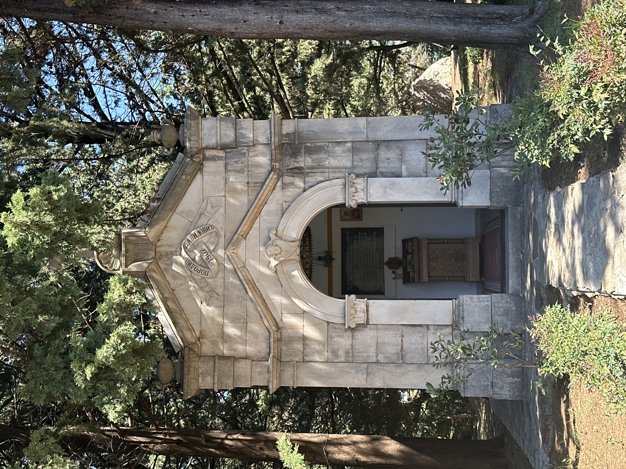 The image shows a small, ornate stone chapel situated outdoors in a wooded area. The chapel is constructed of light-colored stone blocks and features a pedimented facade with decorative carvings. An arched entrance leads into the interior. A stone path leads to the chapel, and there is vegetation, including bushes and trees, surrounding the structure. Inscriptions are visible on the facade of the chapel. The overall impression is of a religious monument or mausoleum set within a natural landscape. The scene appears to be in Boreti, Montenegro.