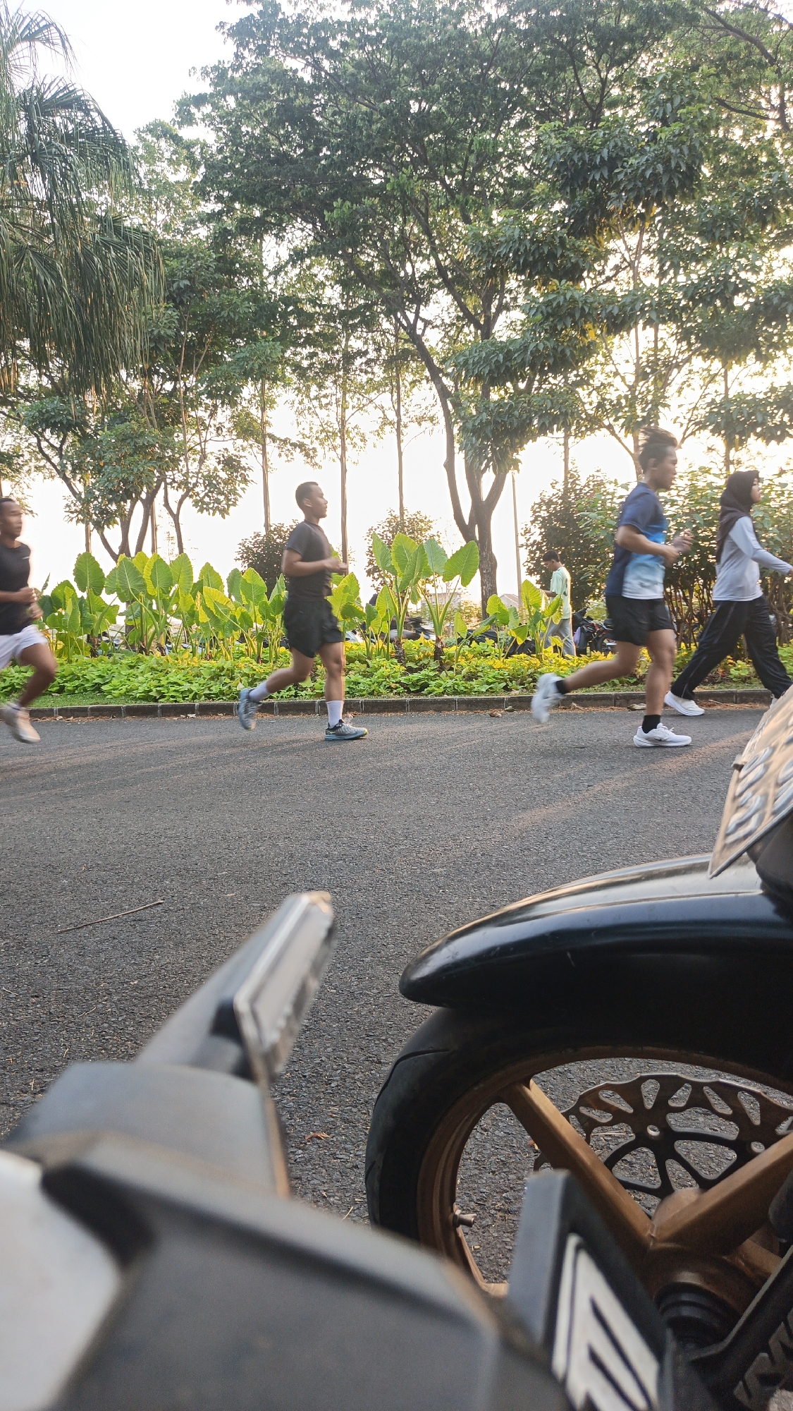 In Bacin, Indonesia, at sunset, several individuals are jogging along an asphalt path bordered by lush greenery and trees. The scene is captured from a low angle, with the foreground dominated by a motorcycle. The sky is a pale yellow, indicating the sun is low on the horizon, casting a warm glow on the environment. The runners are dressed in athletic wear, with one man in a black t-shirt and shorts, another in a blue t-shirt and black shorts, and a woman in a grey hijab and black pants. A fourth person, partially visible in the background, is wearing a light green shirt. The motorcycle in the foreground is black and gold, with its wheel and part of its frame visible. The overall atmosphere suggests a typical evening activity in a park or recreational area.