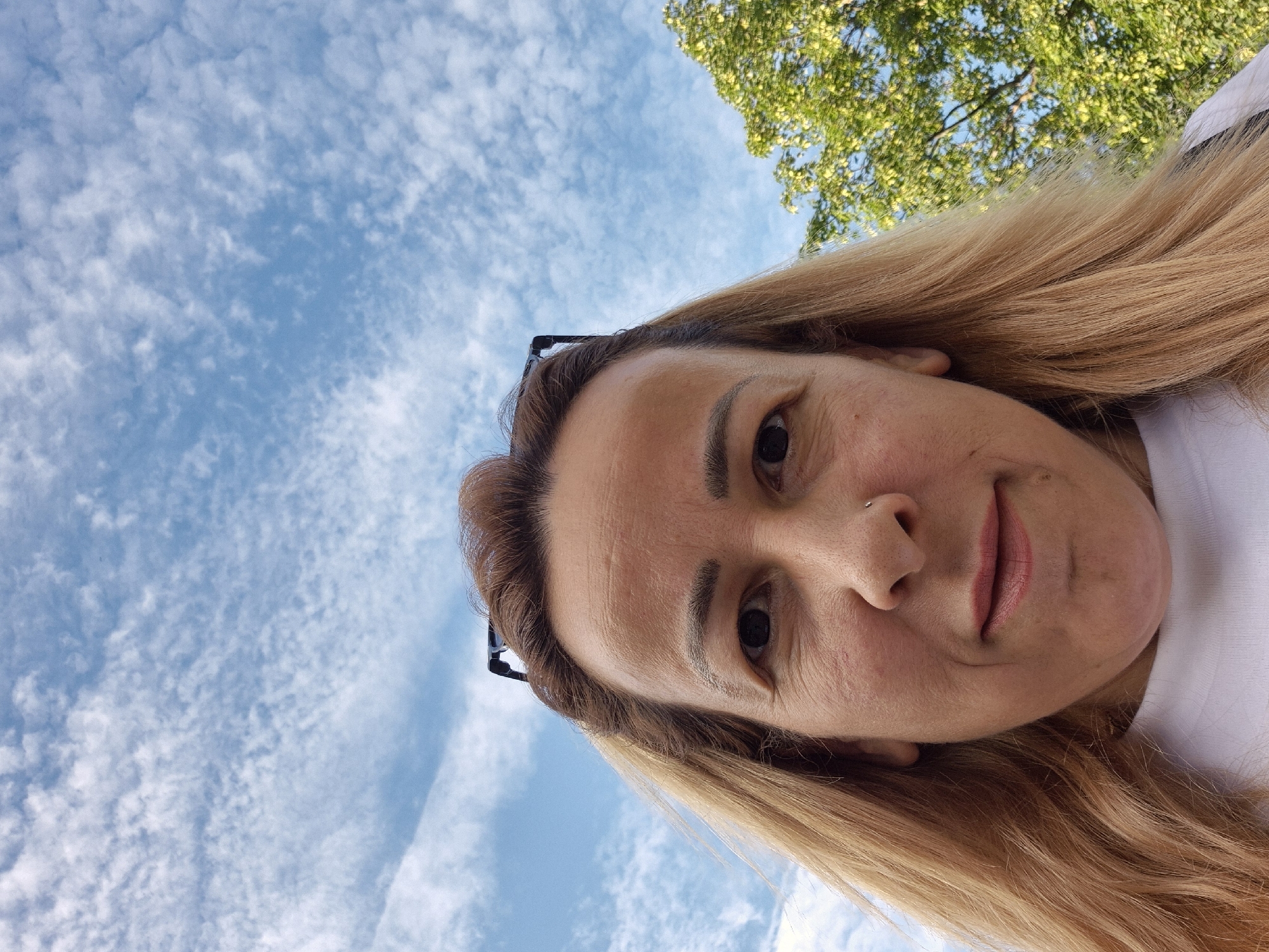 An upward-angled selfie features a woman with shoulder-length blonde hair and a white shirt. She is wearing glasses on top of her head and has a small nose ring. Her expression is neutral, and her lips are slightly parted. The background consists of a bright blue sky with scattered white clouds and a green leafy tree on the right side. The image was taken outdoors, suggesting a daytime setting.