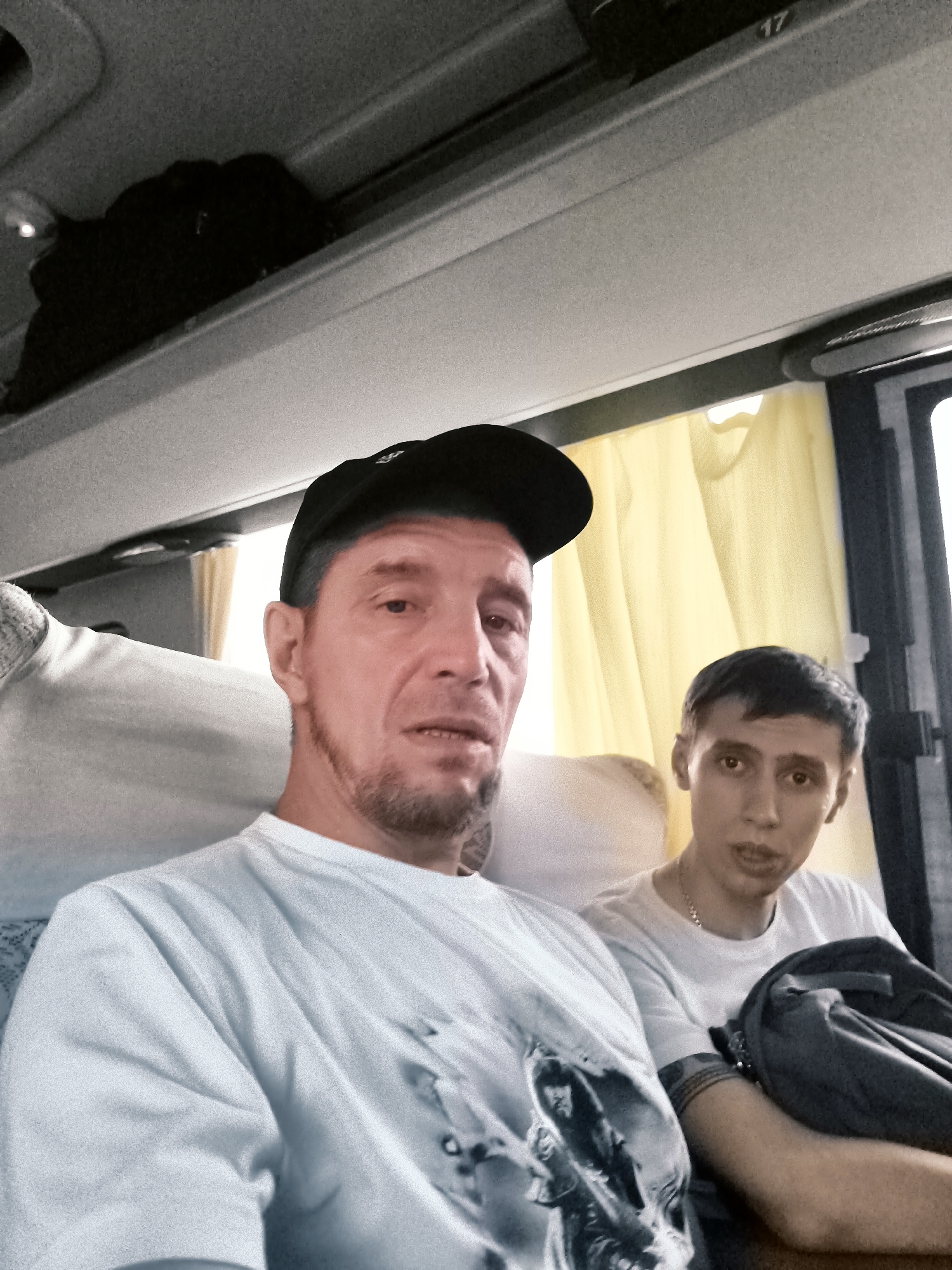 Two males are seated inside a bus with yellow curtains visible in the background. The man in the foreground is wearing a black baseball cap and a white t-shirt with a graphic print. He has stubble on his chin and is looking towards the camera. The man behind him is wearing a white t-shirt and a chain necklace. He is looking to his right. A dark bag is visible on the seat next to him. The environment appears to be a passenger bus. No specific details about a city or location like 果子沟牧场, China are discernible from the image.