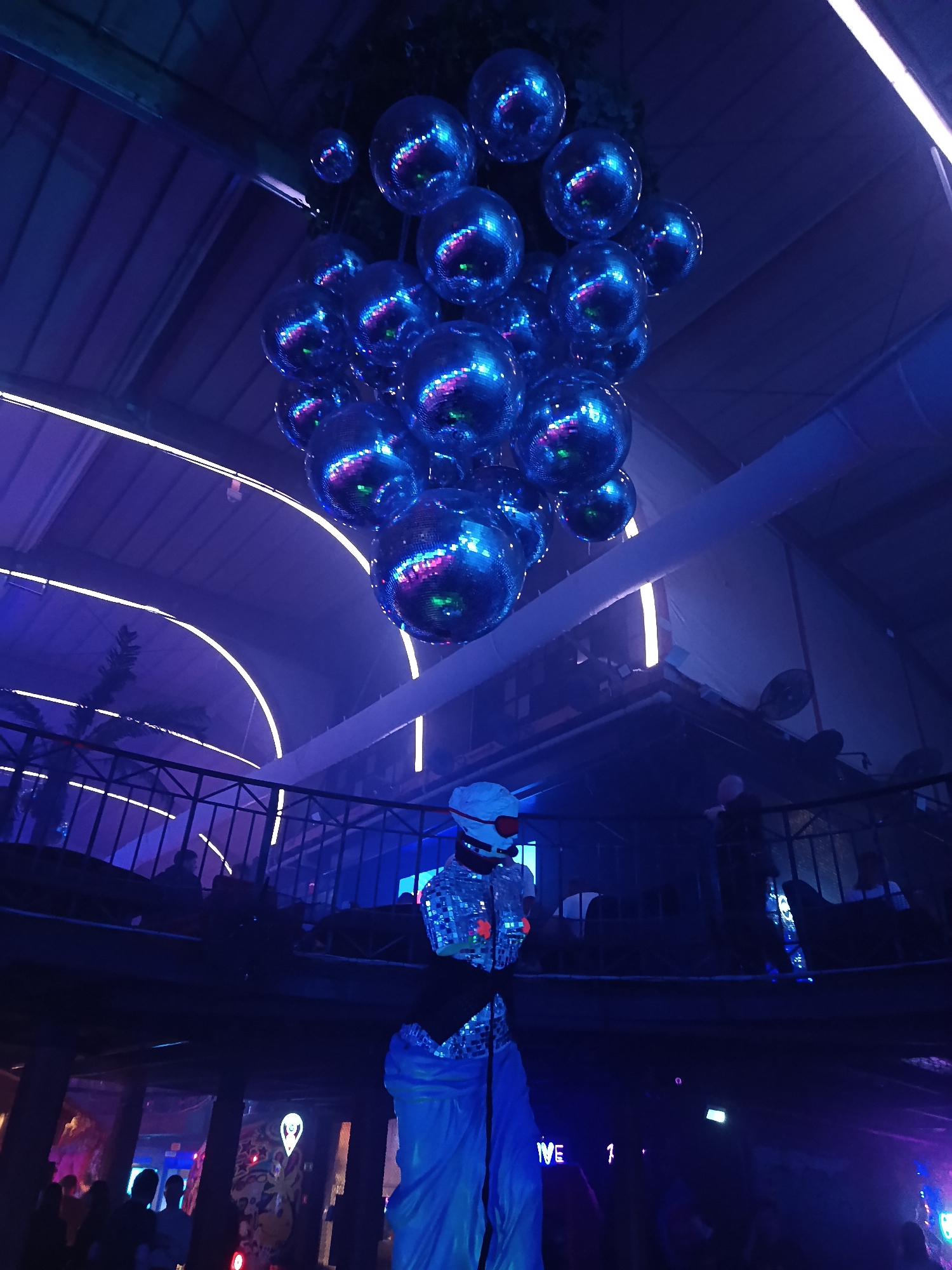 A photograph taken at night indoors features a cluster of disco balls hanging from the ceiling of what appears to be a nightclub. The lighting is predominantly blue, with neon lights visible along the ceiling. Below the disco balls, on a lower level, a figure stands wearing a shiny outfit with a white helmet and what looks like orange lights or reflective material on the chest. In the background, there is a railing, possibly indicating a second level in the venue, where some people are visible. There are people in the foreground as well, though they are mostly silhouetted due to the dim lighting. The overall setting suggests a party or performance event in a nightclub setting. The image does not provide enough detail to definitively place it in Moscow, Russia, but the location could plausibly be an entertainment venue in that city.