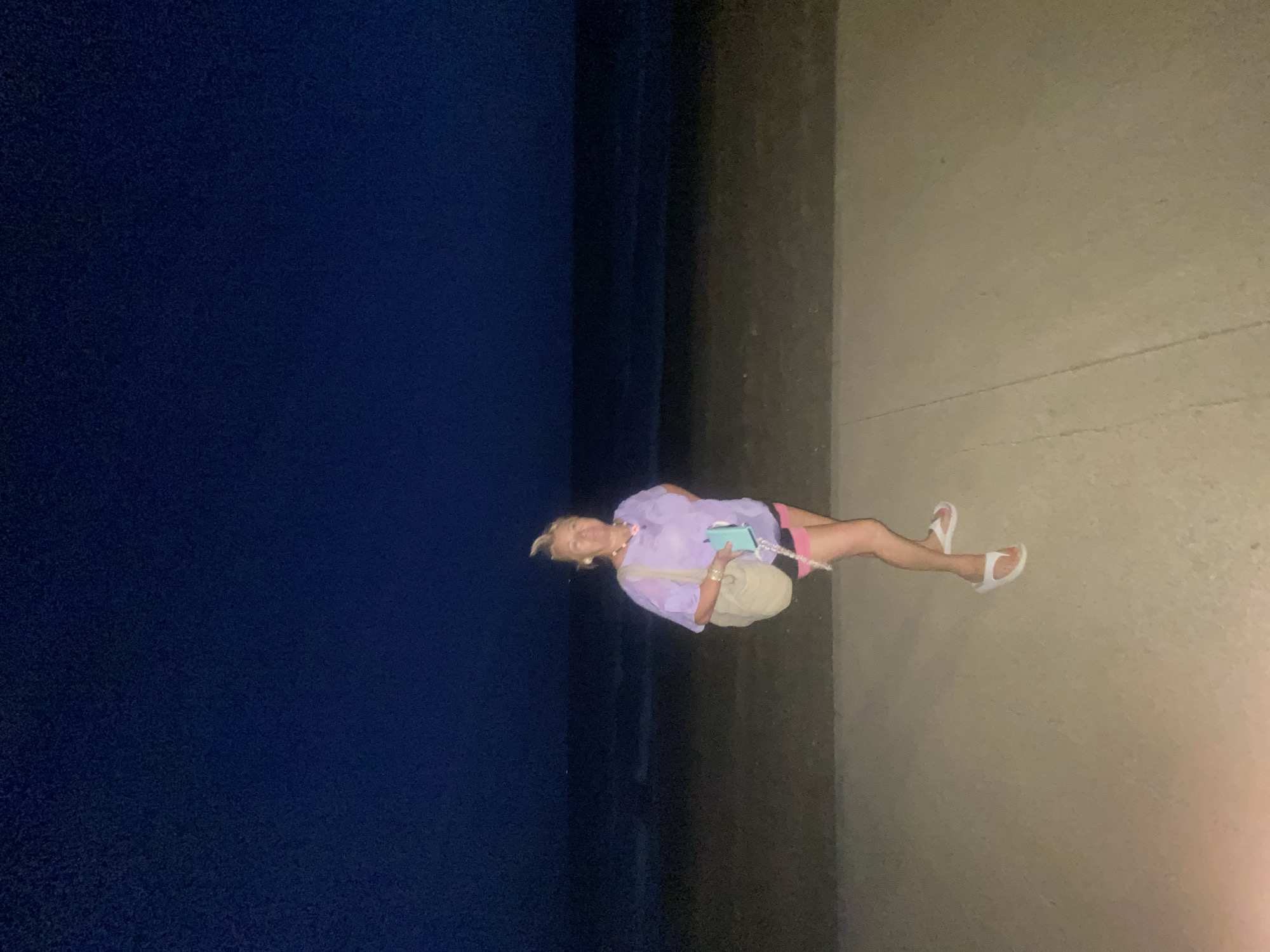 A woman stands on a paved walkway at night. She is wearing a light purple t-shirt, pink and black shorts, and white flip-flops. She is carrying a light-colored bag over her left shoulder and holding a turquoise phone in her right hand. Behind her, the ocean is visible under a dark night sky. The walkway appears to be part of a promenade or beachfront area in Estepona, Spain.
