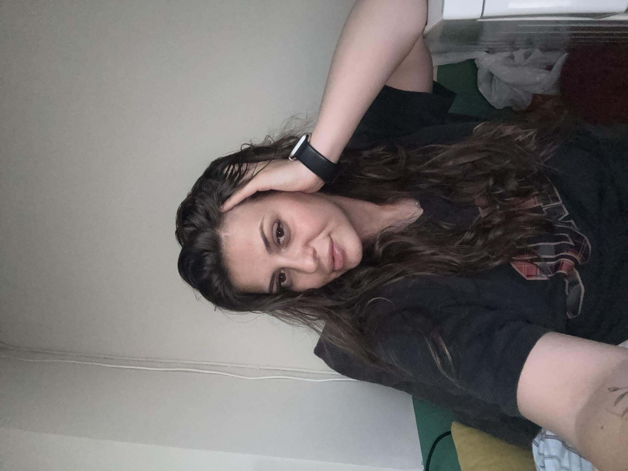 This image, set in the city of Plovdiv, Bulgaria, depicts a female subject, approximately 20-35 years of age, captured in a waist-up, front-facing pose within an indoor environment. She possesses long, dark brown, wavy hair, and a fair complexion. Her dark eyes are directed at the viewer, and her full lips are slightly parted, conveying a neutral to pensive expression. The subject is wearing a dark, short-sleeved t-shirt with a partially visible faded graphic design on the chest and a thin chain necklace with a small pendant. A black Apple Watch with a silver casing is affixed to her left wrist. Her left hand is positioned with fingers touching the left side of her head, appearing to adjust her hair. A faint, indistinct tattoo is visible on her right forearm. The background is a light grey wall, bisected vertically on the left side by a white cable. In the lower left quadrant, a section of green upholstery and a yellow pillow are visible. The lower right quadrant reveals a white object, possibly a piece of furniture or an appliance, next to which a crumpled light-colored plastic bag is present. The lighting is soft and consistent, suggesting an interior domestic setting.