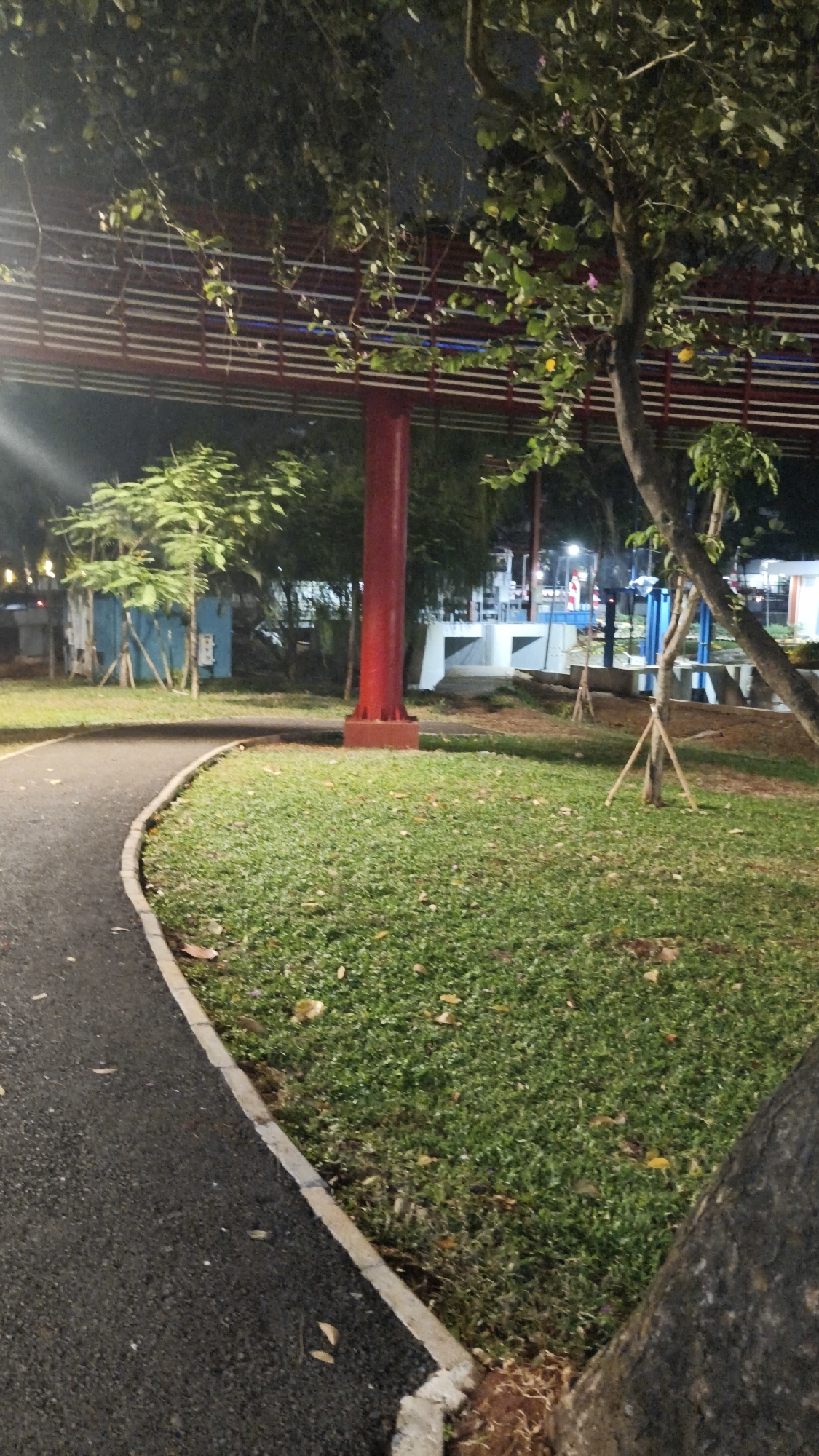 The image shows a park at night in South Jakarta, Indonesia. A paved pathway curves to the left, bordered by a strip of grass. In the background, a tall, bright red pillar supports a red and white striped structure that appears to be an elevated walkway or bridge. Trees with green foliage are scattered throughout the park. The lighting suggests it is nighttime, with some artificial lights visible in the distance, illuminating parts of the park and the walkway. There are no people or animals visible in the scene.