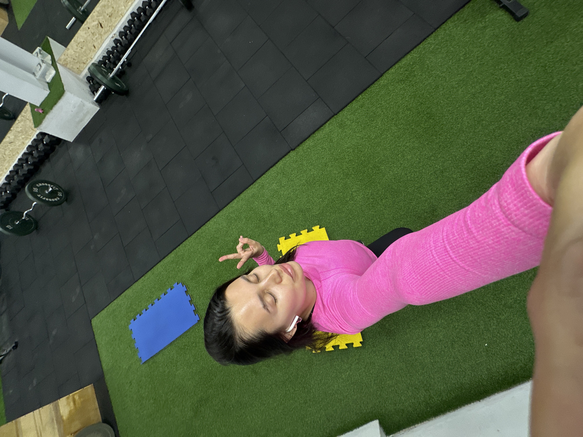 A selfie from an elevated angle captures a person lying on interlocking yellow mats placed on green artificial turf, likely inside a gym. The person, wearing a pink long-sleeve shirt, makes a peace sign with one hand and has an earbud in one ear. A blue interlocking mat is visible nearby. The background shows black interlocking mats and gym equipment, including weights and dumbbell racks. The photo appears to have been taken in Chaikovsky, Russia, although this is not directly discernable from the image itself.