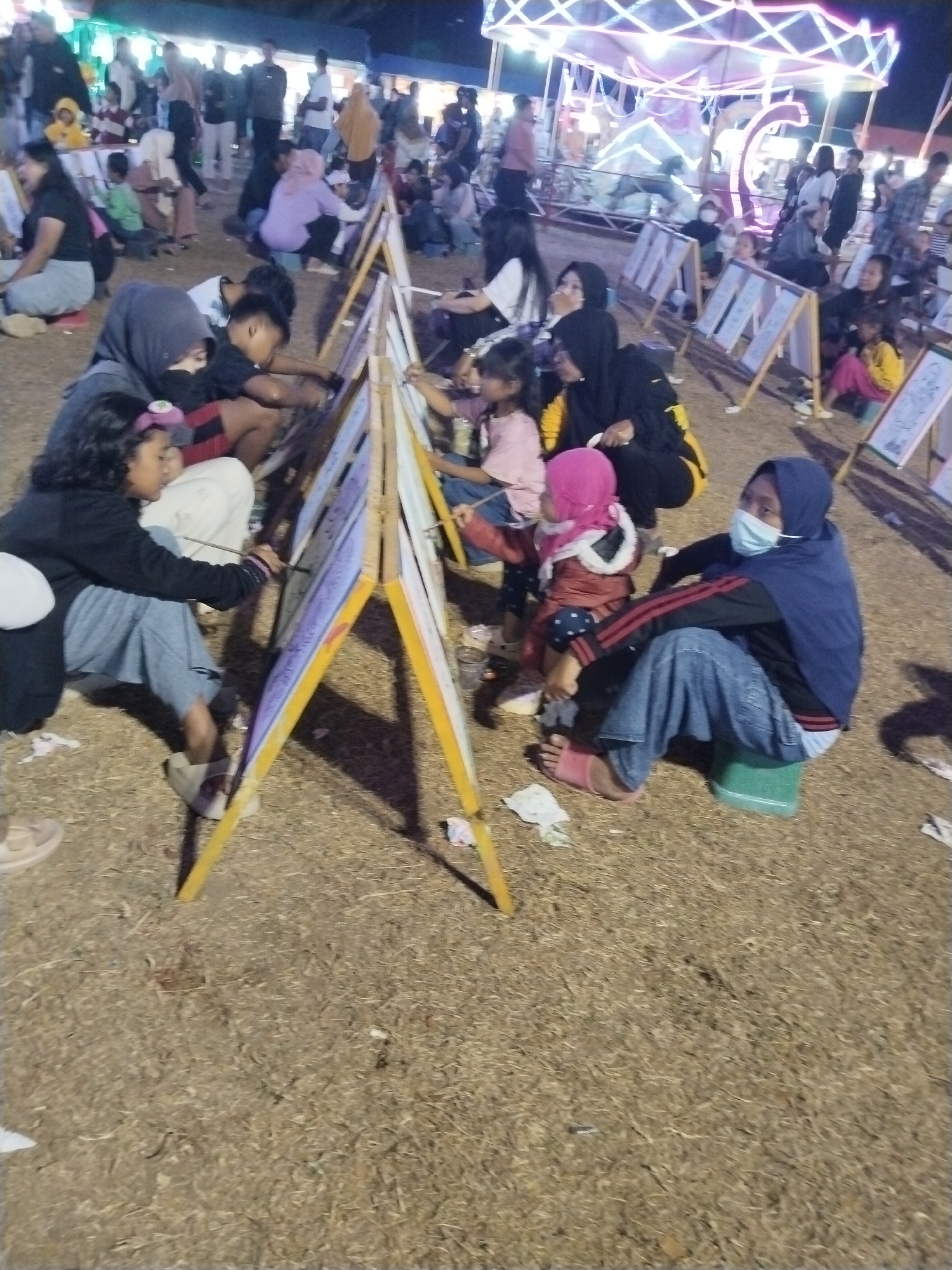 The image depicts an outdoor event, possibly a community gathering or festival, in Tasikmadu, Indonesia. The scene is characterized by a large number of people, including adults and children, engaged in various activities. In the foreground, several children are seated on the ground, intently drawing or painting on canvases supported by wooden easels. One young girl, wearing a black jacket and light blue pants, is actively painting with a brush. Another child, dressed in a pink headscarf and red jacket, is also seated and appears to be participating. To the right, a woman wearing a blue hijab and a light blue face mask is also seated, seemingly observing or waiting. The background reveals a crowded area with more individuals standing and milling about. A brightly lit, illuminated structure, possibly a carnival ride or decorative display, is visible in the upper portion of the image. The ground is covered with dry grass and leaves, suggesting a natural or park-like setting. The lighting indicates it is likely evening or nighttime, with artificial lights from the structures and possibly flash photography illuminating the scene.