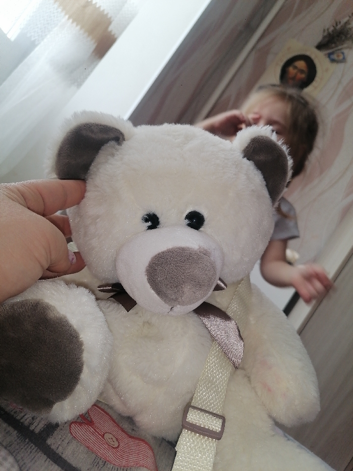This image depicts an indoor scene in Chaikovsky, Russia, dominated by a white plush teddy bear. A light-skinned hand, with visible fingernail polish, holds the top of the bear's left ear. The teddy bear features a white body, gray-brown inner ears and snout, black button eyes, and a small brown bow around its neck. Dark brown patches are present on its paws. A light-colored, woven strap with a plastic buckle runs across the bear's body. In the blurred background, a child with light-colored hair is visible, raising their left hand towards their head. The child appears to be wearing a light gray top. To the left, a window with sheer curtains provides ambient light. The wall behind the child has a light, textured or patterned wallpaper. An oval-shaped religious icon, depicting a bearded figure, is affixed to the wall above the child, with a small sprig of dried lavender or similar plant material attached above the icon. The teddy bear rests on a surface with a visible red and white patterned design, possibly fabric.