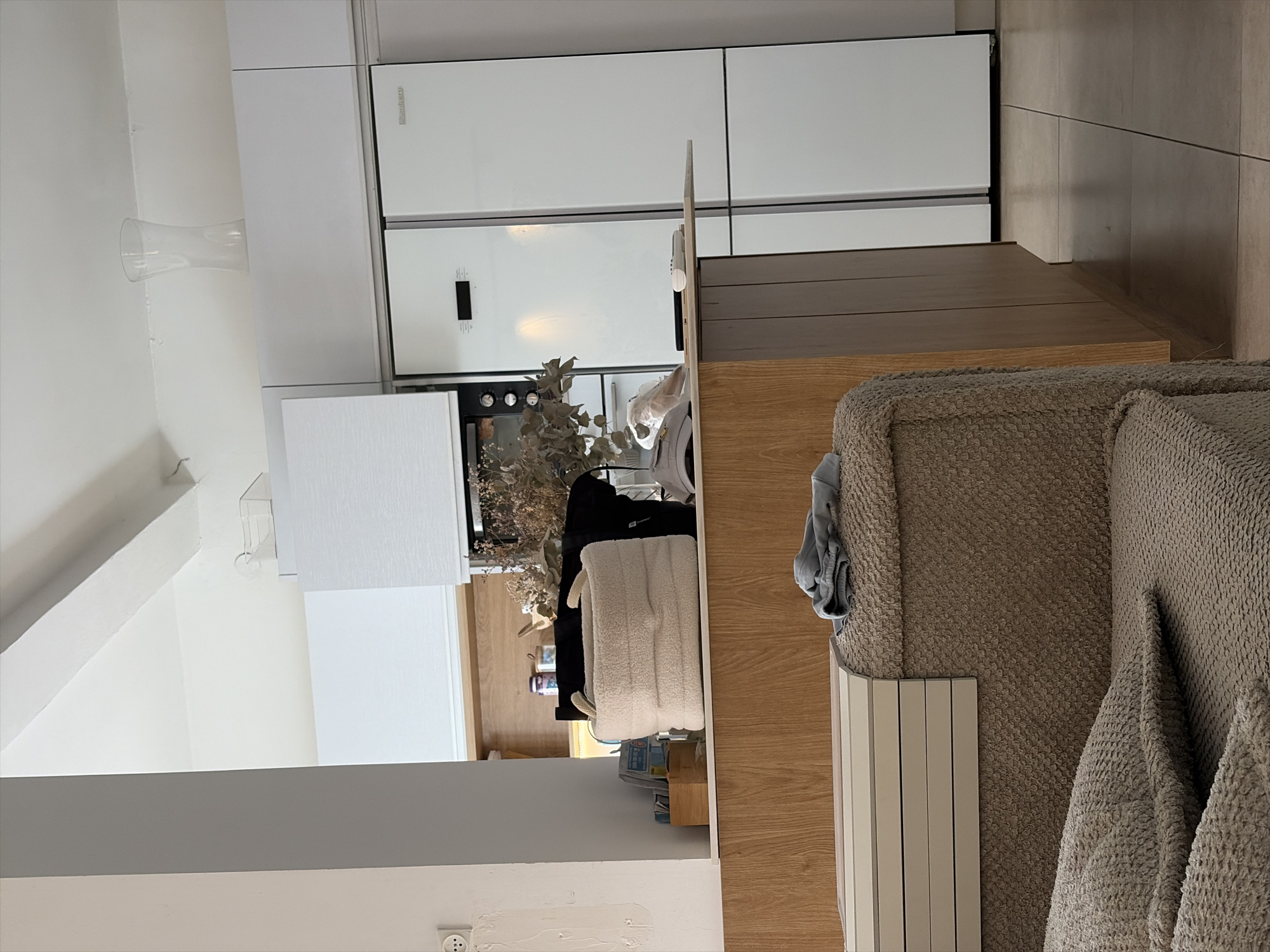 The image depicts an interior scene, likely a kitchen and living area, with a focus on a modern kitchen setup featuring white appliances, including a large refrigerator. In the foreground, a textured brown sofa with pillows is visible, and to the left, a section of a wooden counter or island is present. A light-colored, plush storage basket sits on the counter, alongside some books and a black bag. Above the counter, a vase is visible on a white cabinet. The environment appears to be a contemporary living space. This scene is located in Holon, Israel.