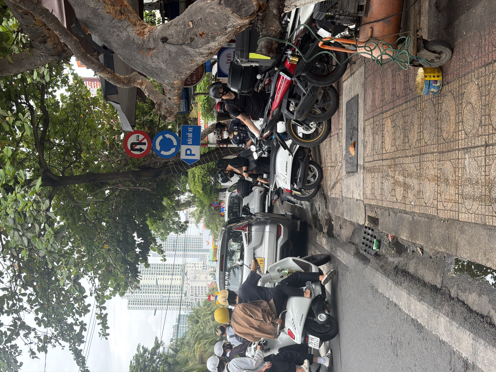 This image captures a vibrant street scene in Nha Trang Ward, Vietnam, during daylight hours, likely overcast. The photo is taken from a slightly elevated perspective, looking down a busy road lined with trees and tall urban buildings in the background.
In the foreground, several individuals are seen on or near motorcycles and scooters. On the immediate right, a scooter carries multiple riders, including one in a yellow helmet and another in a brown jacket, seemingly navigating traffic or preparing to move. Further along the street and parked on the sidewalk, numerous other motorcycles are visible, some with riders still seated or standing nearby, wearing helmets. A silver SUV and a white car are also part of the street traffic further down.
The street itself features a dark asphalt surface with a white lane marking, alongside a patterned sidewalk made of reddish-brown tiles. A drain grate is visible on the sidewalk. Large, mature trees with thick, textured trunks and green foliage line the street, providing shade. In the background, modern, high-rise buildings dominate the skyline, indicating a developed urban area.
Notable details include:
* **Street Signs:** A red circular sign with a crossed-out left-turn arrow over a car, indicating "No left turn for cars," and a blue circular sign with a white arrow forming a circle, suggesting a mandatory direction or roundabout. Another blue rectangular sign with a white "P" and the text "BÃI ĐỖ XE" clearly marks a "Parking lot."
* **Vehicles:** Brands like "LIKEBO" and "AIRBLADE" are visible on some scooters.
* **Other visible text:** A license plate "79-HA 122.47" is seen on a scooter in the foreground. Various other texts and numbers, likely from advertisements, shop signs, or vehicle details, include "0919.357.009," "49 49.7676," "Grob," "02.19," "Con Lầu-C," "DÀN CẦU," "Fm," and "tinh." An old, yellow and blue painted barrel is seen on the sidewalk.