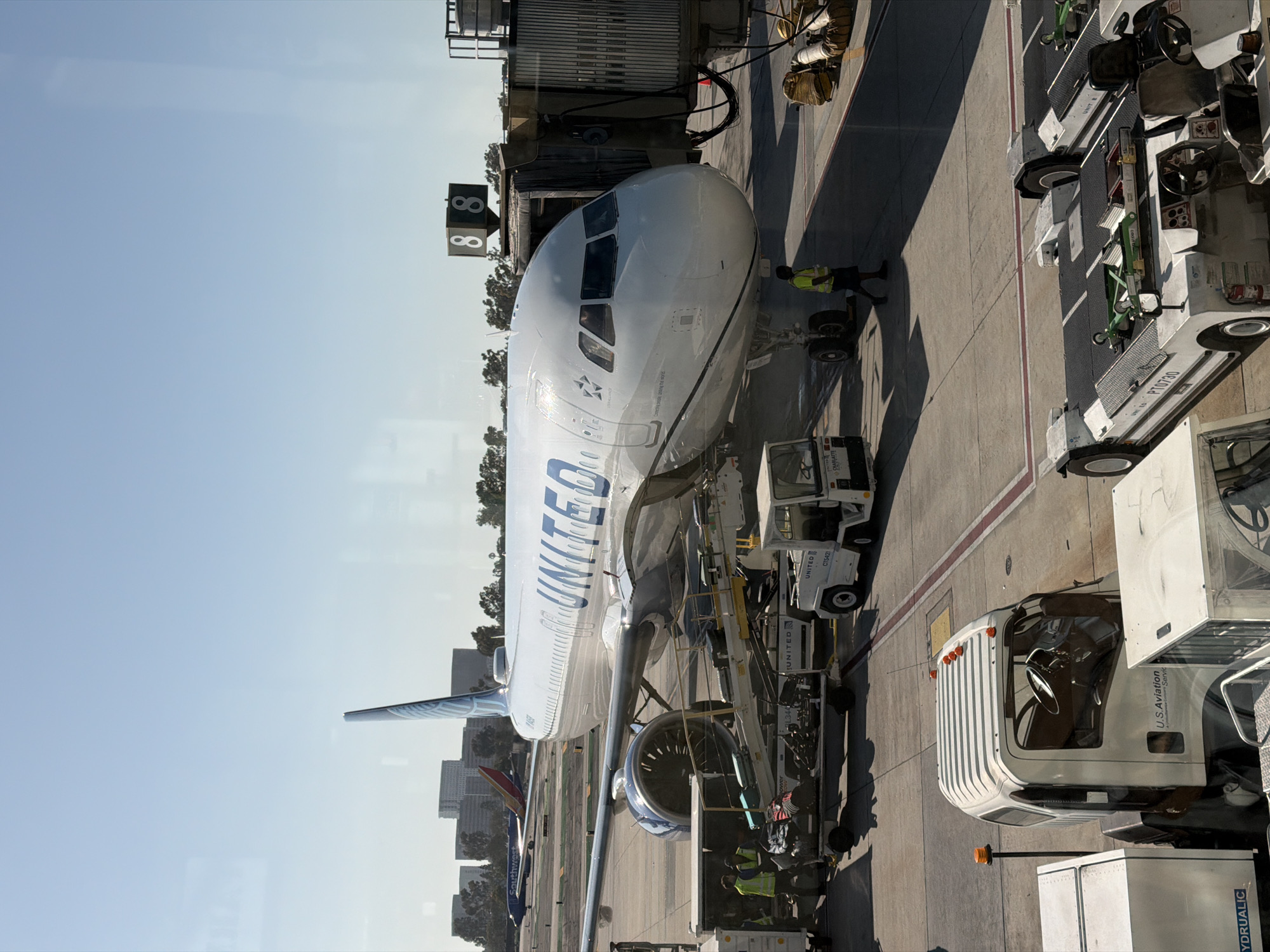 A United Airlines Airbus A320 aircraft is parked at a gate at John Wayne Airport (SNA) in Santa Ana, California, USA. The plane is connected to a jet bridge, and airport personnel are visible around it. Luggage carts, a tug vehicle, and other airport equipment are on the tarmac. In the background, other aircraft and airport buildings are visible. The sky is clear and blue.