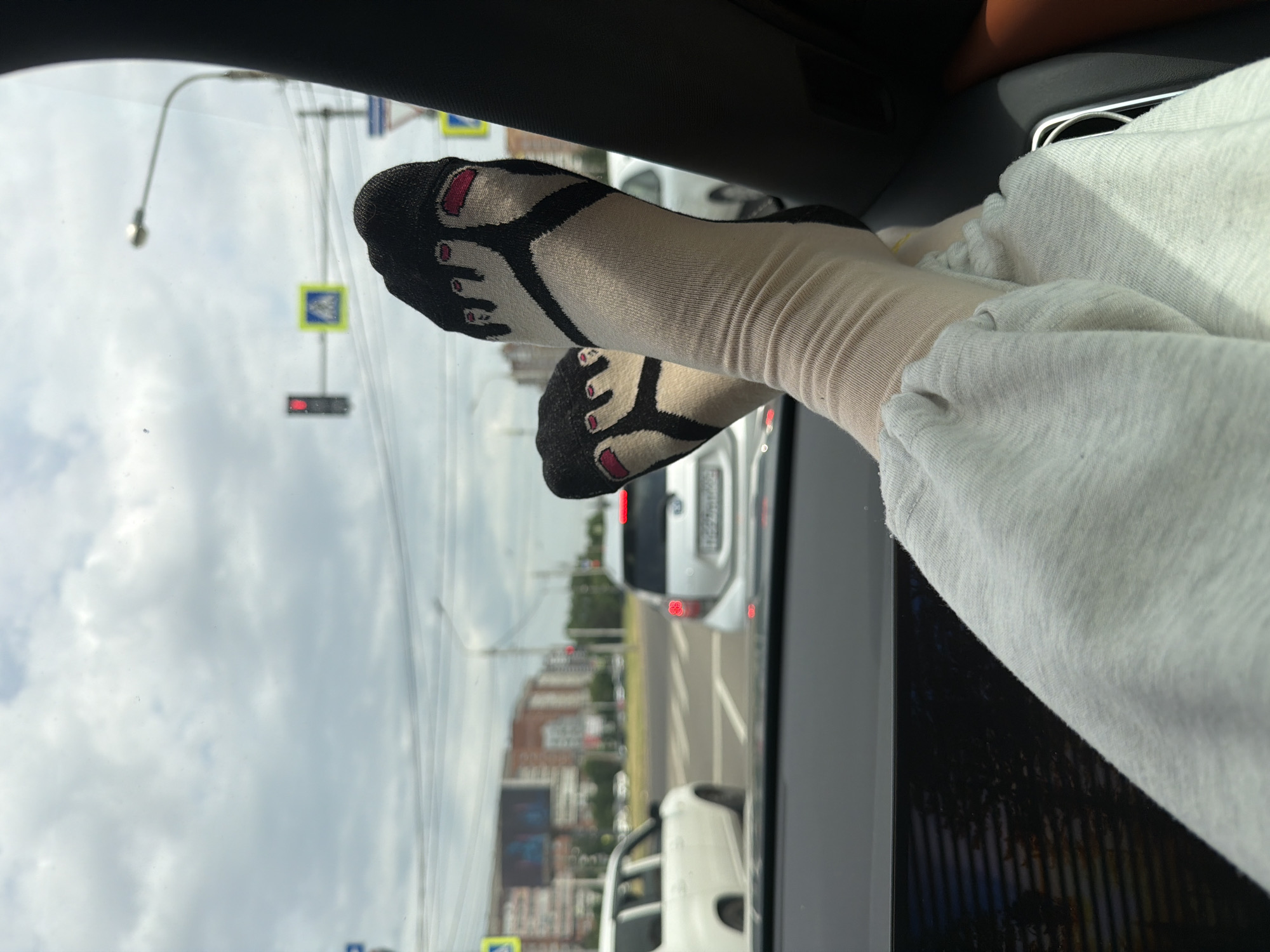 The image shows a pair of feet wearing novelty socks with a cartoon design resembling toes. The socks are light-colored with black and red details. The feet are resting against a car's interior, with a light-colored garment, likely sweatpants, also visible. The scene appears to be captured from inside a vehicle, looking out through the window.
Outside the car, a street scene unfolds. There are traffic lights, pedestrian crossing signs with yellow backgrounds and blue symbols, and several cars on the road, including white ones. Buildings, possibly apartment blocks, are visible in the background, with some trees and greenery. The sky is overcast with clouds. The presence of specific traffic signs and the architectural style of the buildings suggest the location is Krasnoyarsk, Russia.
The overall mood seems relaxed, as indicated by the casual posture of the feet and the comfortable clothing. The time of day appears to be daytime, given the natural light and the illuminated traffic lights. The weather seems to be cloudy. There is no visible text in the image.