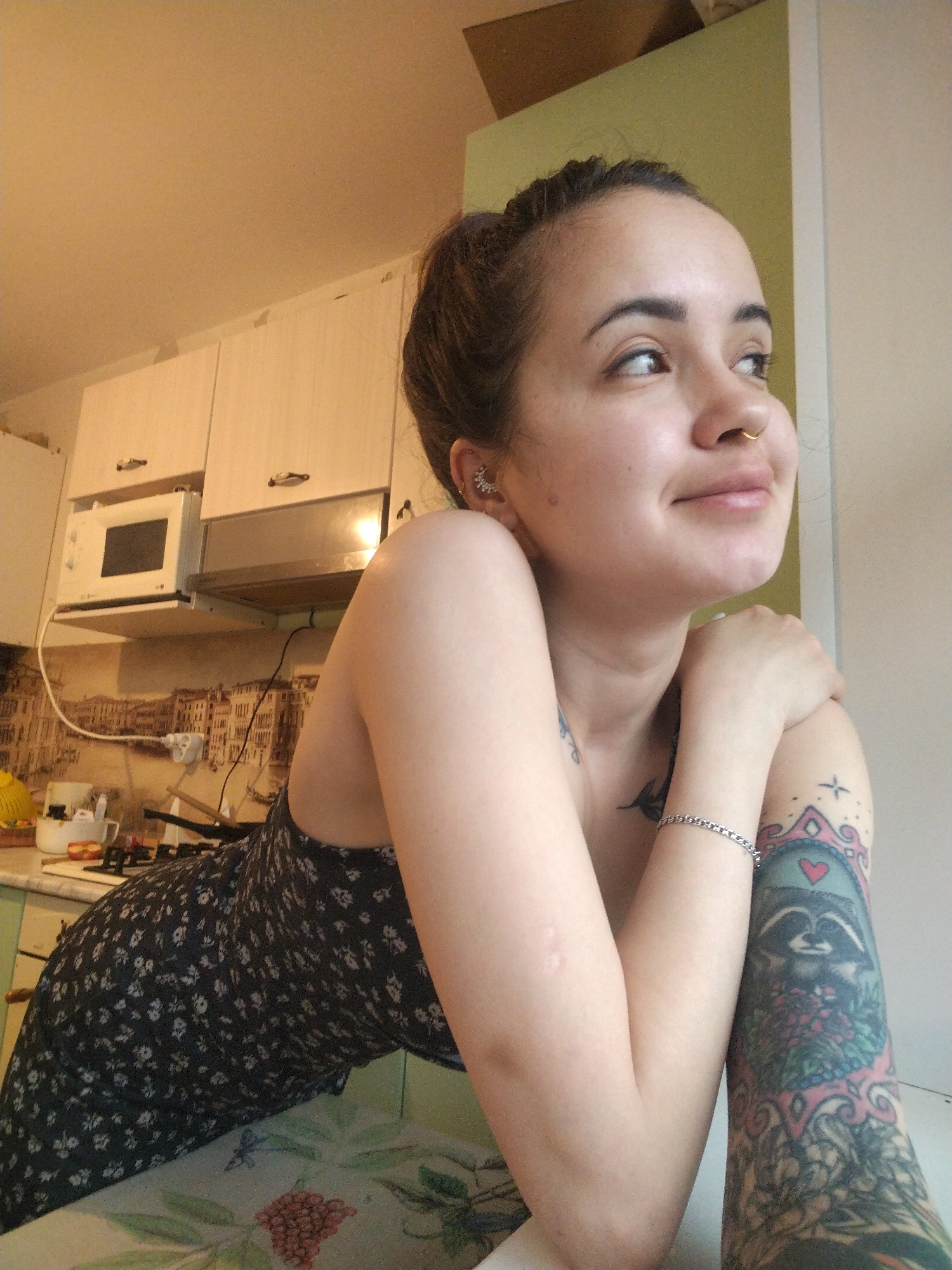 A young woman poses indoors, likely in a residential kitchen. She is fair-skinned with dark hair pulled back in a bun, and has piercings in her ear and nose. She wears a dark floral dress and a silver bracelet on her wrist. Visible tattoos adorn her chest and right arm, featuring a raccoon within a decorative frame. She is leaning on a table, arms crossed. Behind her, kitchen cabinets and a microwave are visible, along with a patterned backsplash. The environment suggests a domestic setting. The location is implied, but not directly evident. Noviy is not visibly a location in this image.