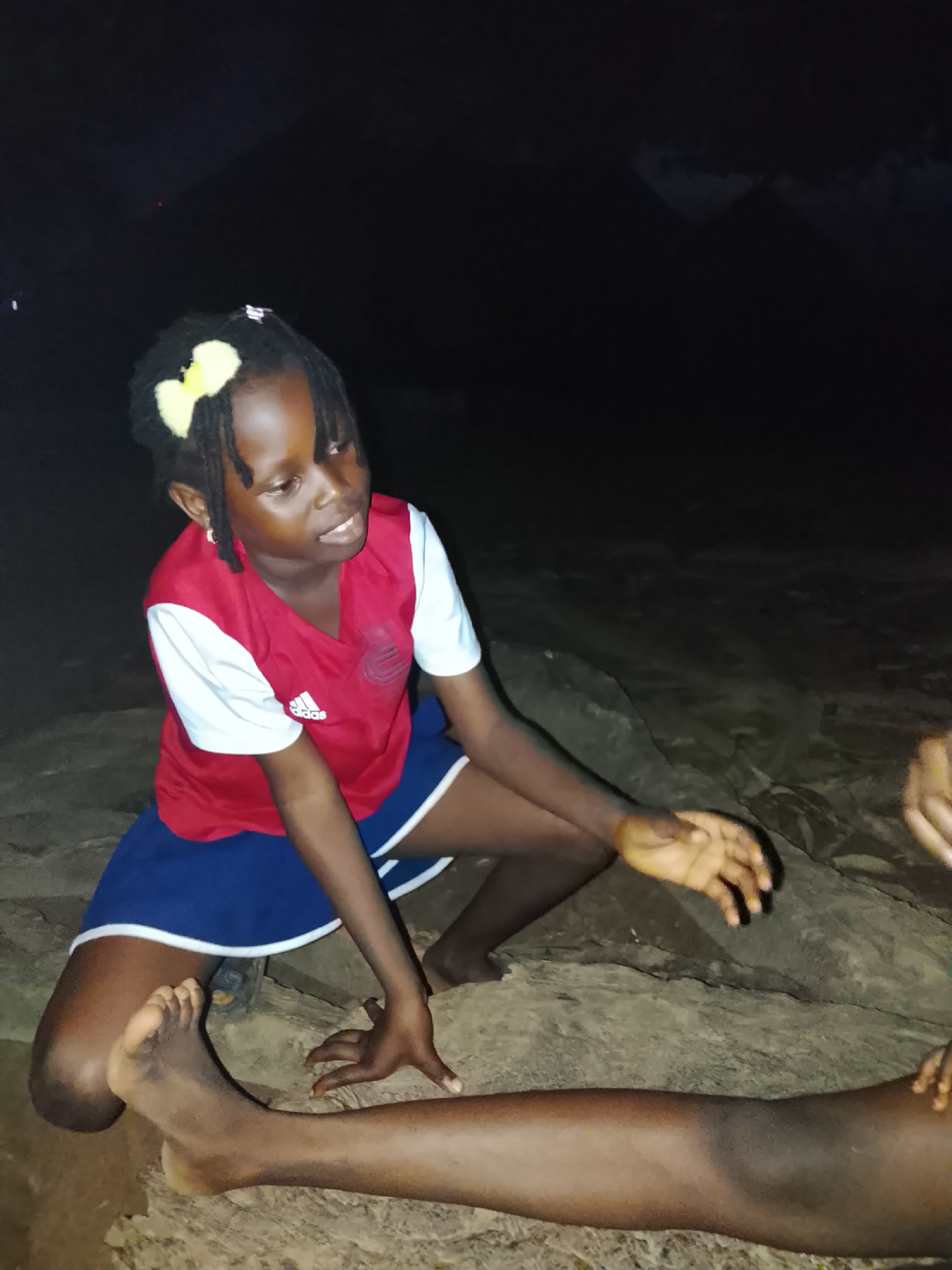 In Akwanga, Nigeria, a young girl with dark skin and braided hair adorned with a yellow bow sits on rocks at night. She is wearing a red and white t-shirt with blue shorts. Her right hand rests on the rock, while her left arm is bent at the elbow, with her hand extended forward as if reaching for something. Another person's legs are visible in the foreground, lying horizontally across the frame. The background is dark, suggesting it is nighttime, with faint shadows of what appear to be mountains in the distance. The lighting is dim, casting a warm glow on the subjects.