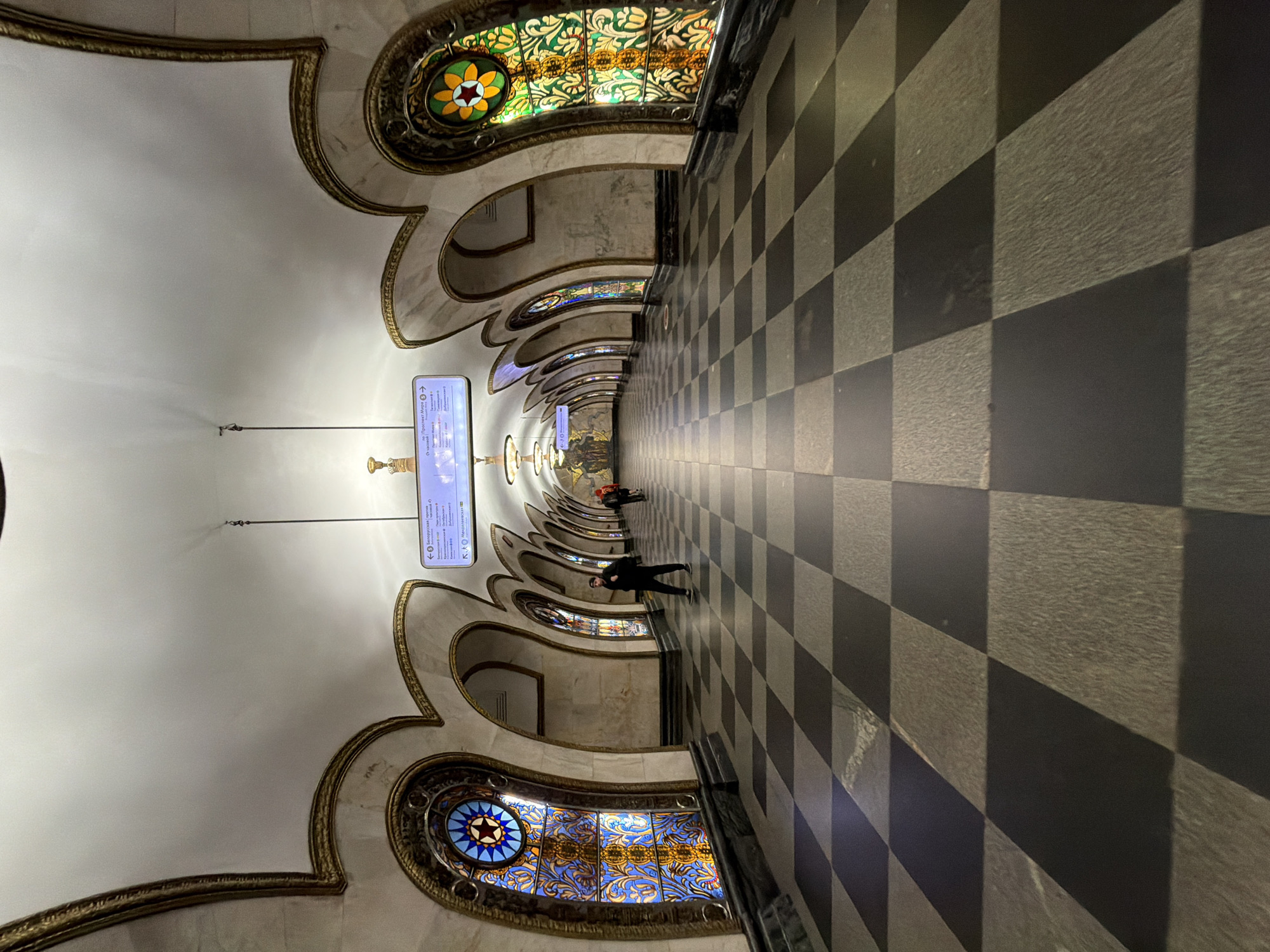 This image depicts the Mayakovskaya station in the Moscow Metro, Russia. The station features ornate, vaulted ceilings with gilded edges and stained glass panels. A tiled floor, patterned in black and gray squares, extends into the distance. Two people are walking through the station, and signs with the Moscow Metro logo are visible.