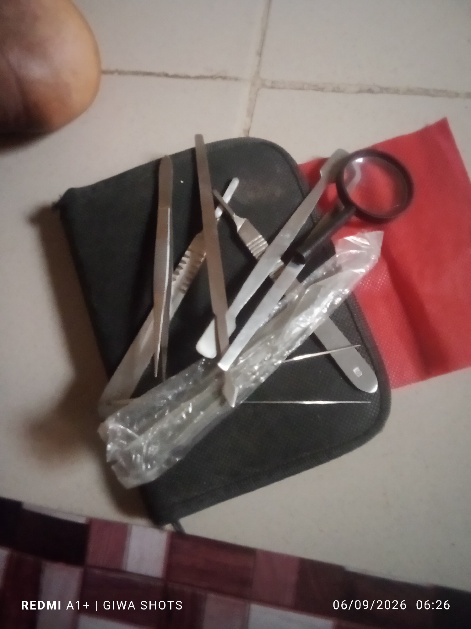 The image shows a collection of medical or dissection tools laid out on a black case. The tools appear to be made of metal and include tweezers, scalpels, and a magnifying glass. A red cloth is visible behind the tools. The scene is likely indoors, with a tiled floor visible in the background. The lighting is somewhat dim, casting shadows on the tools and case. The bottom of the image contains text indicating the phone model and date.