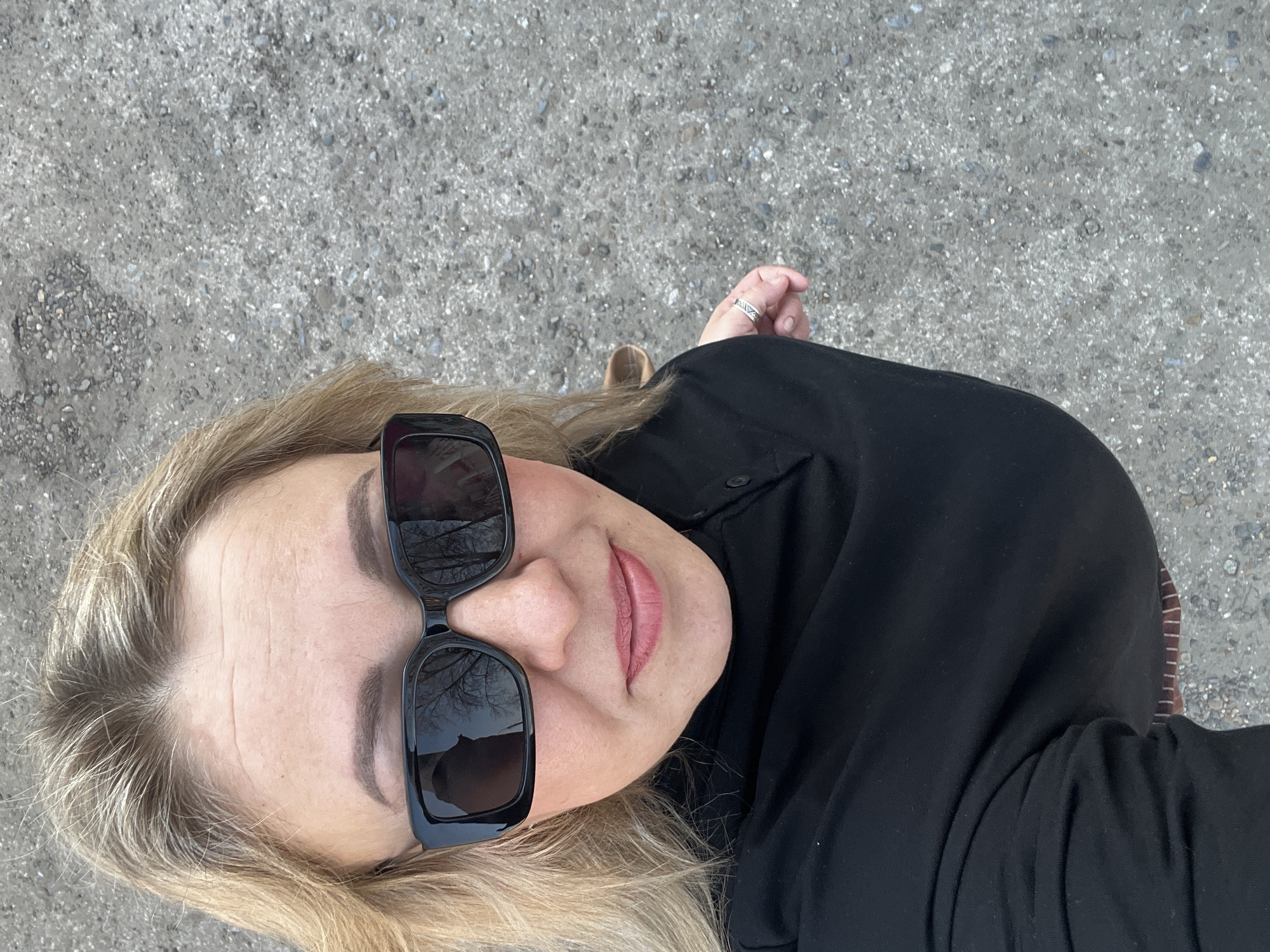 A top-down, selfie-style shot shows a blonde-haired woman with sunglasses standing on a paved surface, likely a street or sidewalk. The glasses reflect bare tree branches, suggesting an outdoor location possibly in late fall or winter. She is wearing a black shirt. Her hand is visible with a silver ring on her finger. The background is a gray, textured concrete or asphalt surface. Given the prompt, the location may be inferred to be in or around Chaikovsky, Russia, but this is not visually verifiable.
