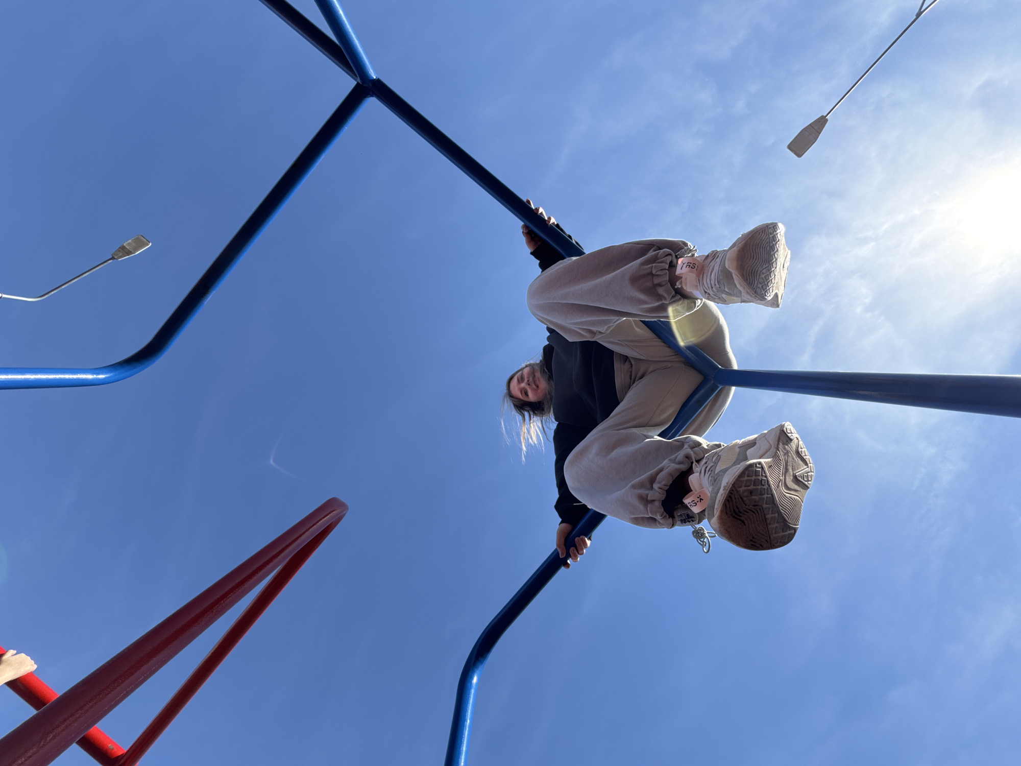 A low-angle shot shows a person on a playground structure against a bright blue sky. The person is wearing a dark top, light-colored sweatpants, and sneakers. The person is hanging upside down from a blue metal playground structure. A red metal structure is visible in the lower left corner of the frame. A streetlight is visible in the background. The location is Горный, Russia.
