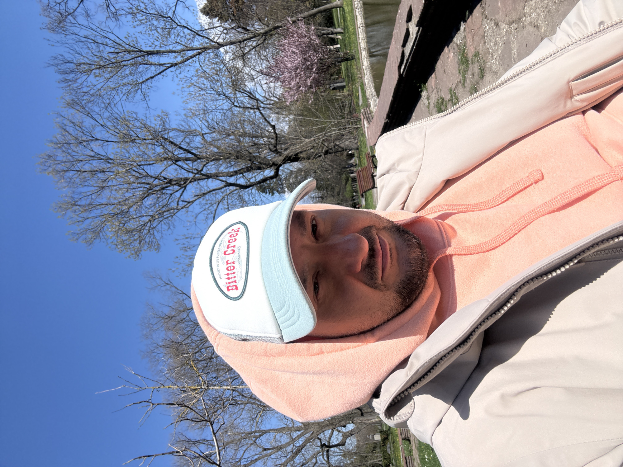 A man is pictured outside on a sunny day. He is wearing a white baseball cap with the logo "Bitter Creek, Wyoming", a light peach-colored hoodie, and a light grey zip-up jacket. Trees, some bare and some with pink blossoms, are visible in the background, along with a stone wall and water. The scene is likely in a park or green space. The photograph shows details suggesting it was taken near a waterfront area within the city of Sofia, Bulgaria.
