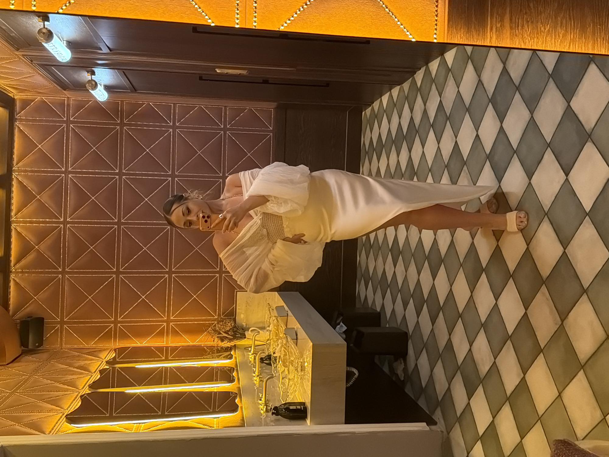 A full-body mirror selfie captures a woman posing in what appears to be a luxurious restroom or powder room. The woman, with dark hair pulled back, is wearing an off-the-shoulder, cream-colored formal dress with voluminous, sheer puffed sleeves and a fitted bodice. The lower part of the dress is a flowing, satin-like skirt featuring a high slit on her left leg. She wears simple nude-colored slide sandals. Her right hand holds a light-cased smartphone up to take the photo, partially obscuring her face, though a ring is visible on her right hand.
The environment is richly decorated. The walls behind her, visible in the mirror's reflection, are covered with textured panels in a warm, gold-bronze hue, featuring a raised, stitched diamond pattern. Along the top of the wall on the left, concealed lighting casts a soft, warm glow. Below this, a white marble or stone vanity features two ornate, clear glass basin sinks with gold-toned, wall-mounted faucets. A dark soap dispenser and a small, dried floral arrangement are placed on the vanity. Beneath the vanity, dark support structures or bins are visible.
To the right of the woman, the actual mirror's frame is wide and dark, possibly wood, and adorned with vertical strips of small, warm, twinkling lights. Reflected in this mirror frame are dark wood paneled walls, possibly doors, fitted with two cylindrical, modern wall sconces that emit a cool, bluish-white light. The floor throughout the scene is covered in a checkered diamond pattern of light and dark grey or black and white tiles. A small, dark speaker is mounted on the wall in the upper left of the reflected room.