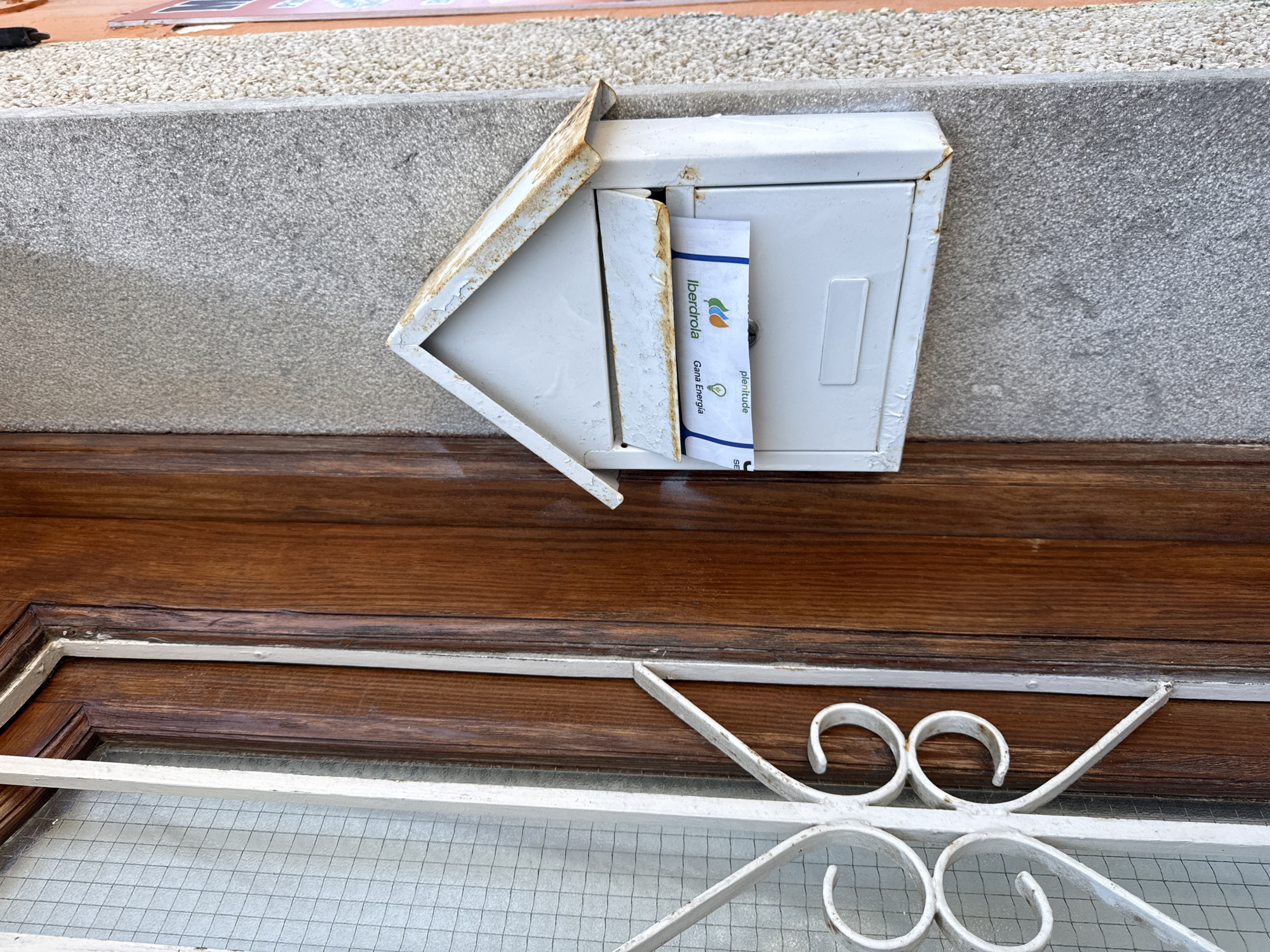 The image shows a white mailbox on a grey, textured wall next to a wooden door with a white wrought iron grate. A piece of mail with "Iberdrola Gana Energía" printed on it is sticking out of the mailbox. The environment suggests an outdoor setting, possibly the entrance to a residential building. The scene is static and appears to be a typical residential exterior. The specific location in Blanes, Spain, cannot be determined from the visual information alone.