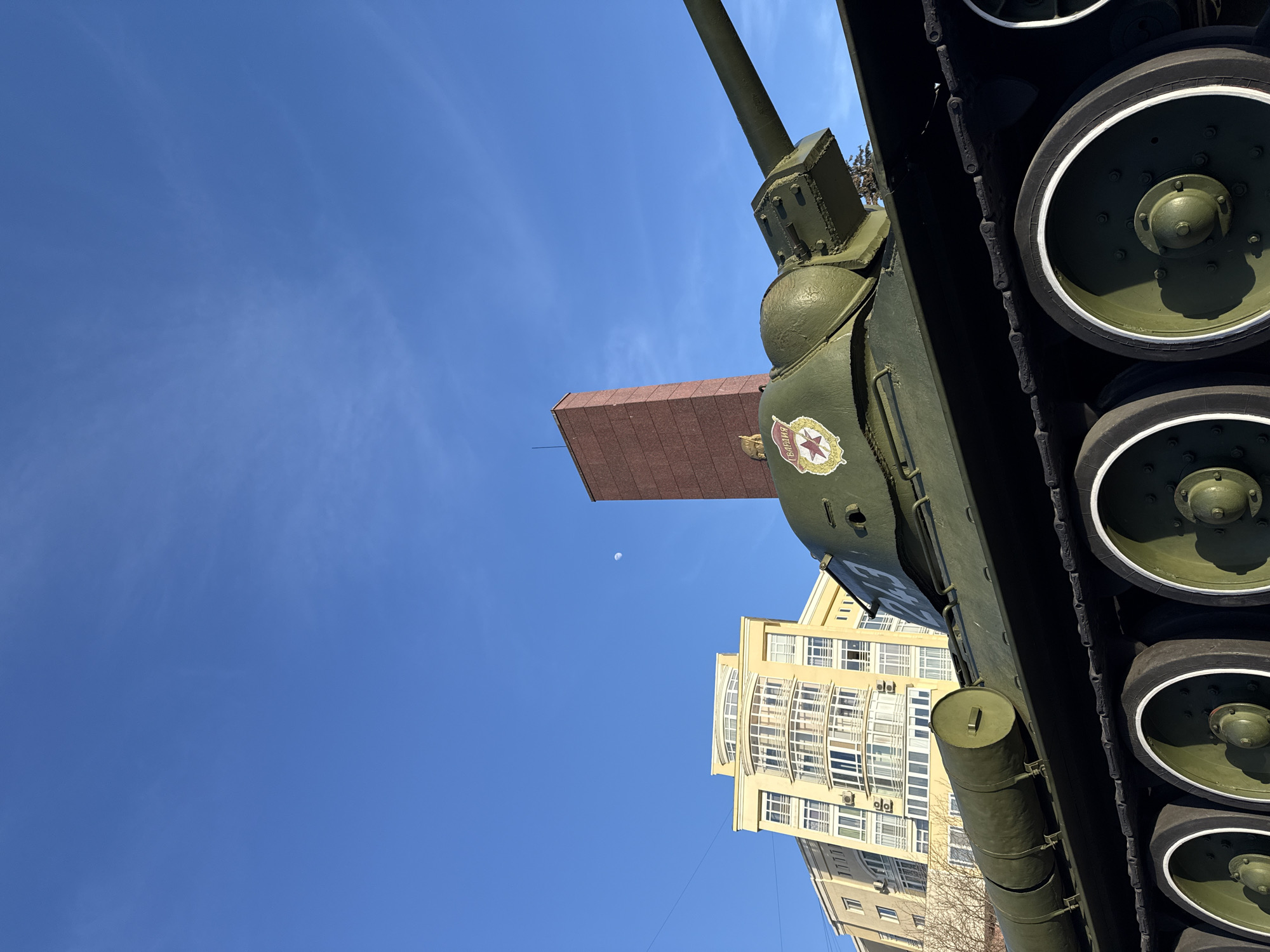 The image presents an outdoor, urban scene dominated by a static, olive green T-34-85 medium tank monument. The tank is positioned in the foreground, viewed from its left side and slightly front, featuring its visible road wheels with dark green hubs and white rims, and a section of its dark metal tracks. Two cylindrical fuel tanks are mounted horizontally on the tank's hull. The turret displays a red star emblem encircled by a wreath, with the Cyrillic word "ГВАРДИЯ" (Guardia) positioned above it, and the number "243" on its side. The tank's main gun barrel extends towards the right of the frame. In the background, a multi-story, light yellow/beige building with numerous windows and curved balconies is visible to the left. To the right, behind the tank, a tall, slender, reddish-brown rectangular column-like structure rises. The sky is clear and blue with minimal wispy clouds, and a faint crescent moon is discernible in the upper center. The scene is bathed in bright daylight. This tank monument is located on Komsomolsky Prospekt, Perm, Russia.