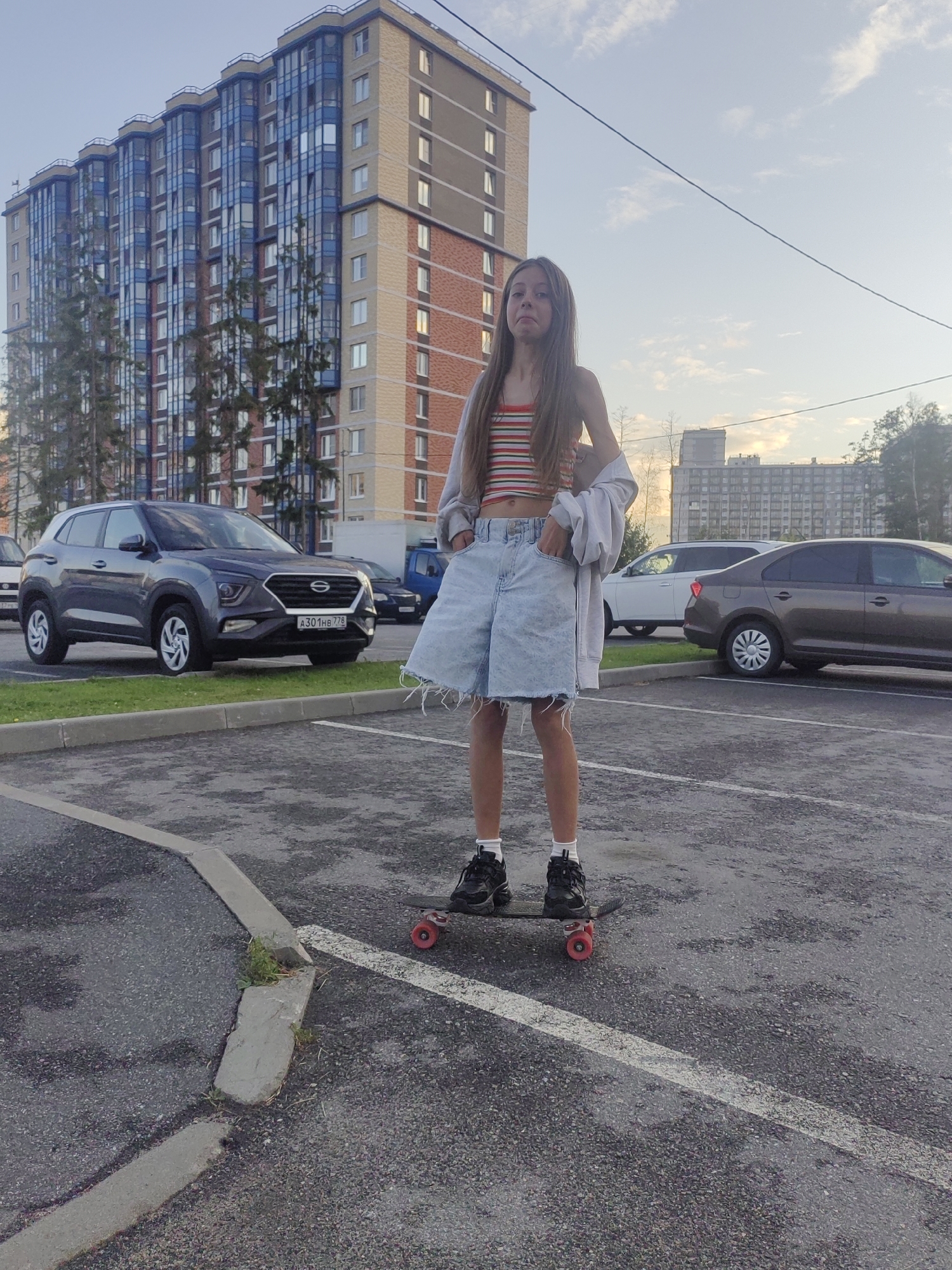 A young female with long brown hair stands on a skateboard in a parking lot. She is wearing a striped tank top, denim shorts, and sneakers. Behind her, a modern apartment building with blue and brown facades is visible, along with several cars and trees. The sky is overcast, suggesting it is either early morning or late evening. The scene is consistent with a residential area in Saint Petersburg, Russia, where such apartment complexes are common.