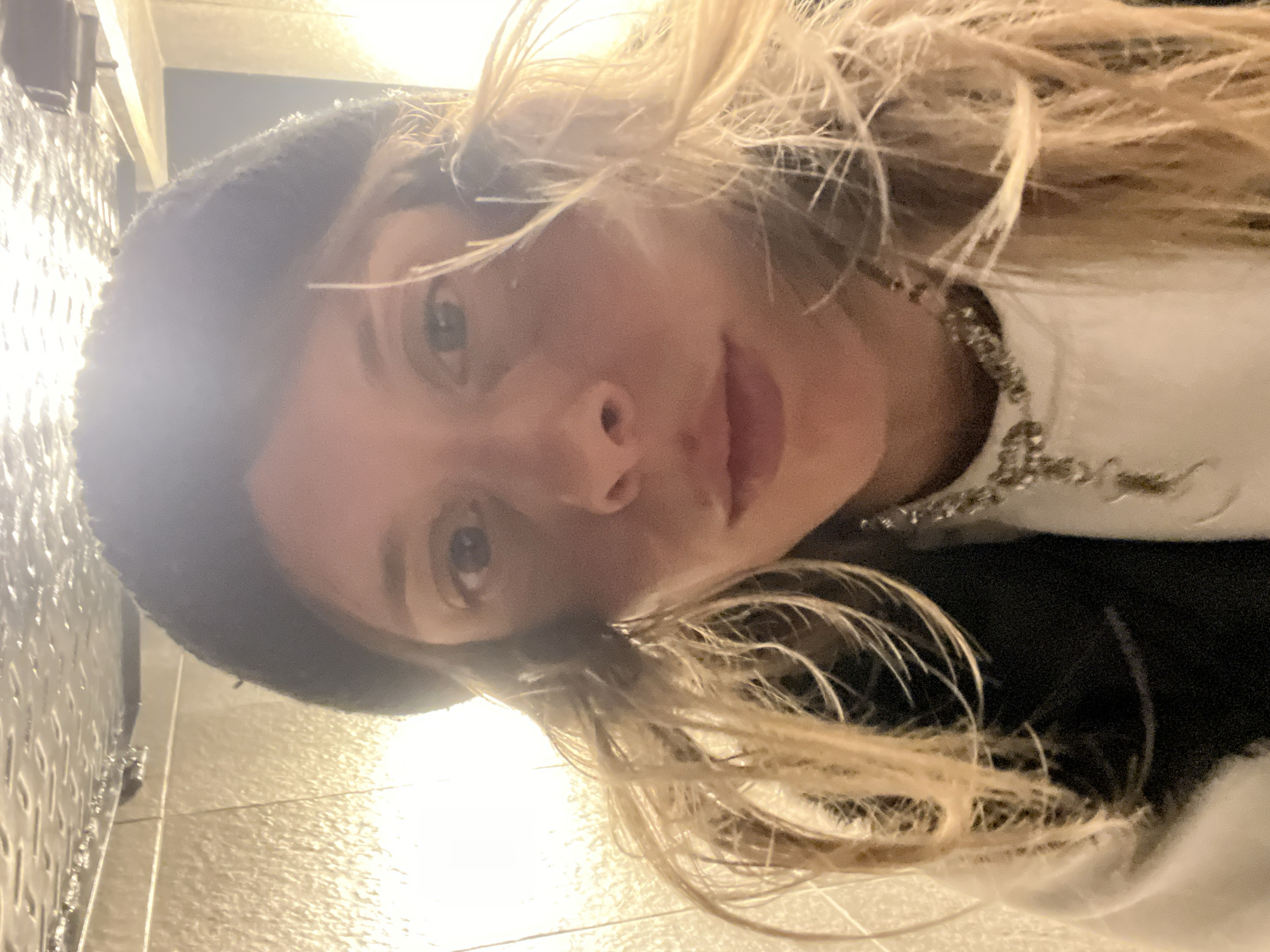 The image is a selfie of a woman with blonde hair, blue eyes, and fair skin, wearing a black beret. She is wearing a white top with what appears to be a silver necklace or embellishment. The background shows a tiled wall. The image appears to have been taken indoors, possibly in a restroom or similar space with tiled walls. There's no direct indication of Buenos Aires, Argentina.