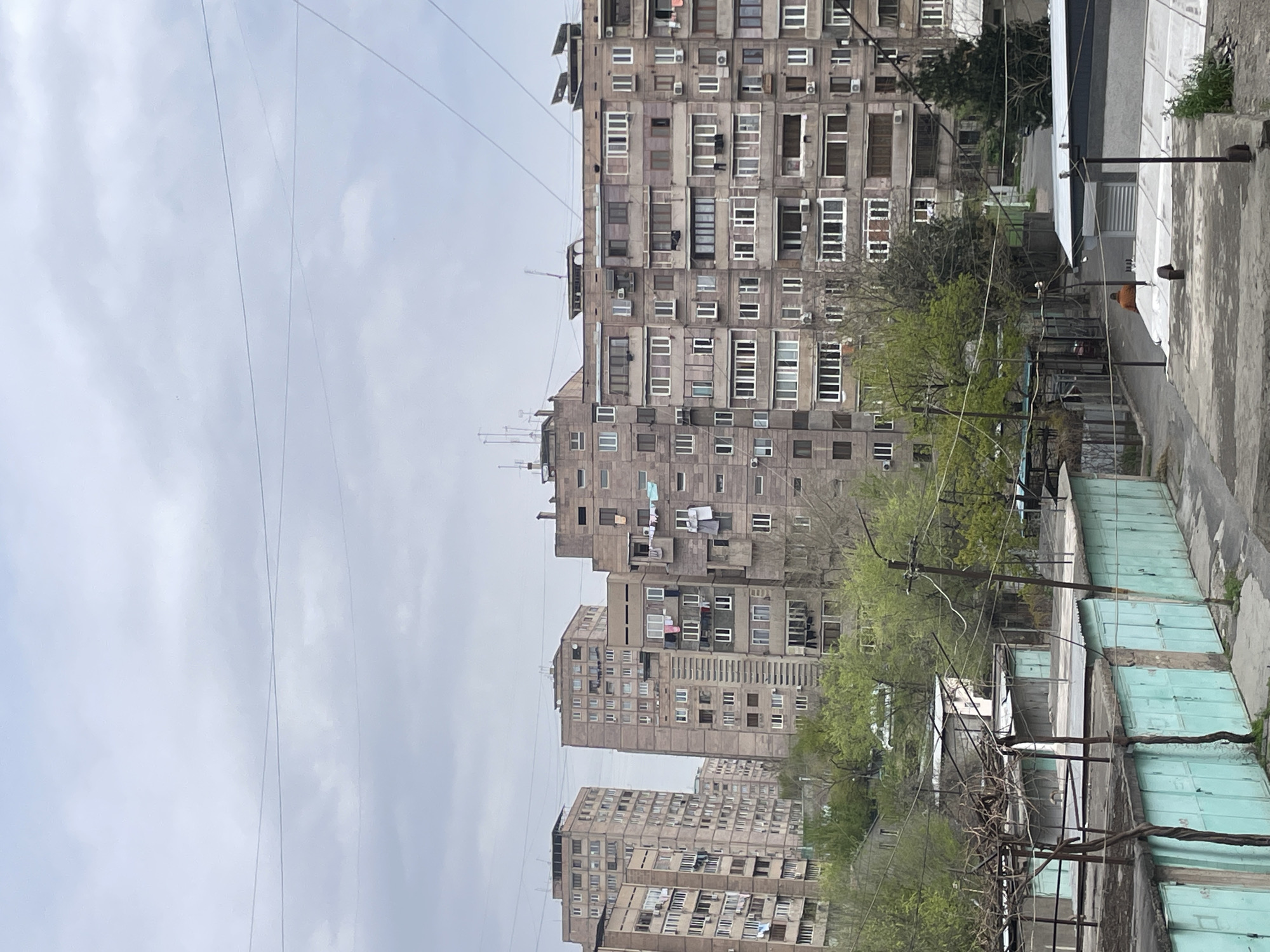 The image shows a view in Yerevan, Armenia, dominated by Soviet-era apartment buildings constructed of grey concrete. The buildings are tall and repetitive, with many windows and balconies. In the foreground, there are garages, also constructed of concrete, painted in a light blue-green color. Trees with new spring foliage are interspersed between the buildings and garages. Power lines crisscross the sky. The sky is overcast and grey. There are clothes hanging out on one of the balconies.