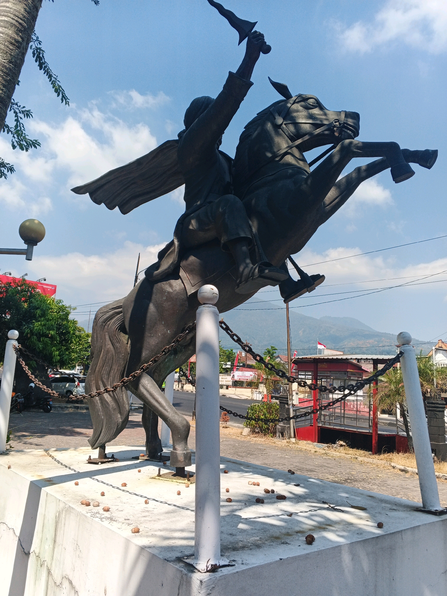 The image depicts a bronze statue of a man on horseback in a dynamic pose, with the horse rearing up on its hind legs. The man is wearing traditional attire and appears to be raising a sword or dagger in his right hand. A cape or cloak flows behind him, suggesting movement. The statue is situated on a concrete pedestal, which is surrounded by a chain-link fence and white posts. Small, nut-like objects are scattered on the base of the pedestal.
In the background, there are trees, buildings, a street with vehicles, and distant mountains. A red building with glass windows and a white awning is visible to the right of the statue. An Indonesian flag, red and white, is flying from a pole in front of the red building. The sky is blue with scattered white clouds, and the overall lighting suggests a sunny day.
Based on visual cues and common knowledge of Indonesian landmarks, this statue is likely located in Ungaran, Indonesia. Specifically, it is the General Gatot Subroto statue, a significant monument in the city, commemorating the national hero. The statue is situated near the Ungaran city square and administrative center.