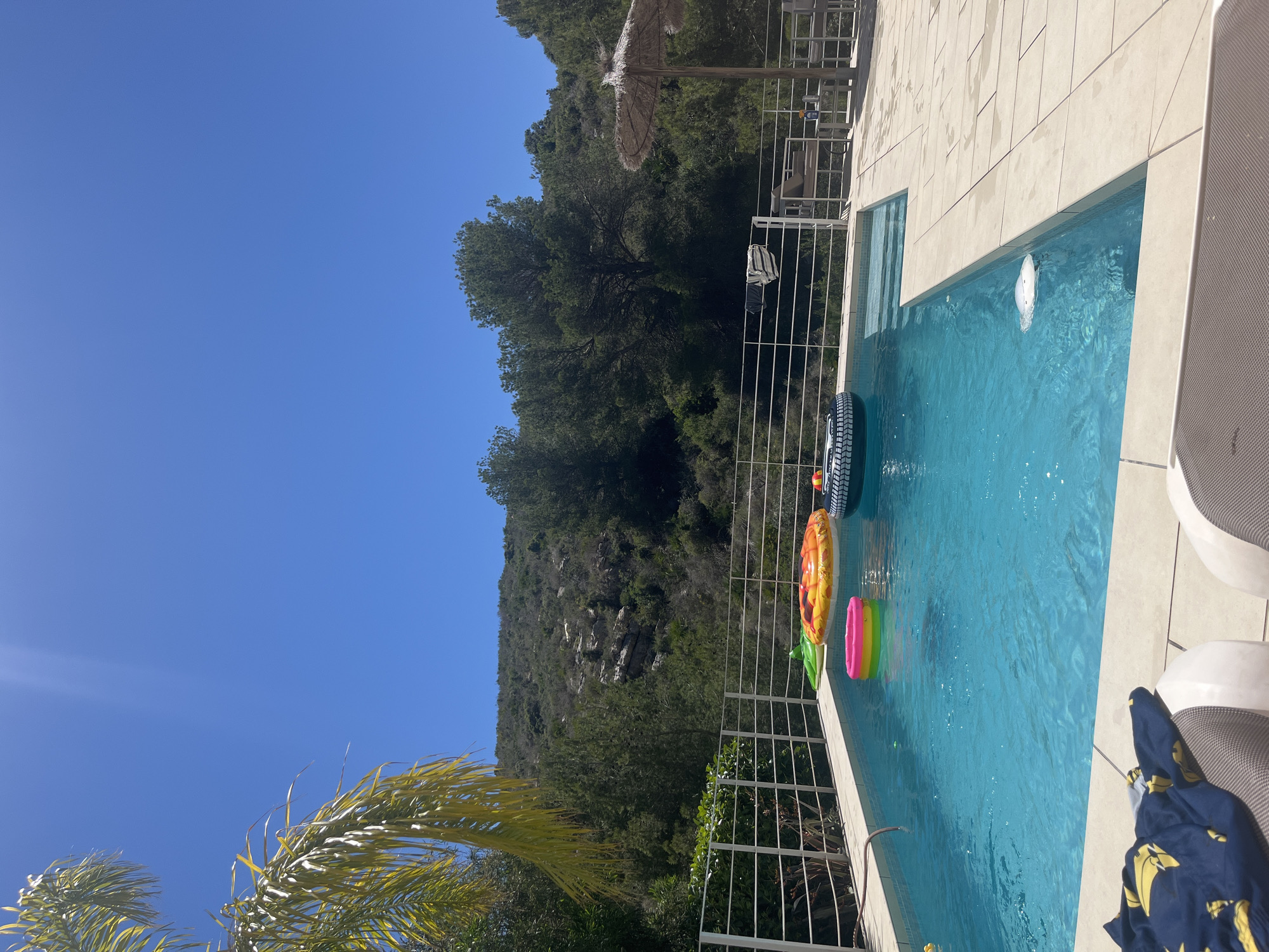 The image depicts a rectangular swimming pool in a residential setting, likely in el Poble Nou de Benitatxell/Benitachell, Spain, given the local flora and architectural styles commonly found in the area. The pool is filled with clear, turquoise water, containing several inflatable pool toys including a black and white tire, a pineapple and a multi-colored ring. The pool is surrounded by light-colored tiles and a white metal fence that separates the pool area from the natural landscape. The background reveals a lush green hillside with trees and rocky outcroppings, typical of the Mediterranean region. Above the hill, a clear blue sky dominates the upper left portion of the image. A straw sun umbrella and a metal chair are seen in the background, suggesting a leisure or vacation environment. In the foreground, part of a lounge chair and a blue shirt with yellow motifs on it can be seen.