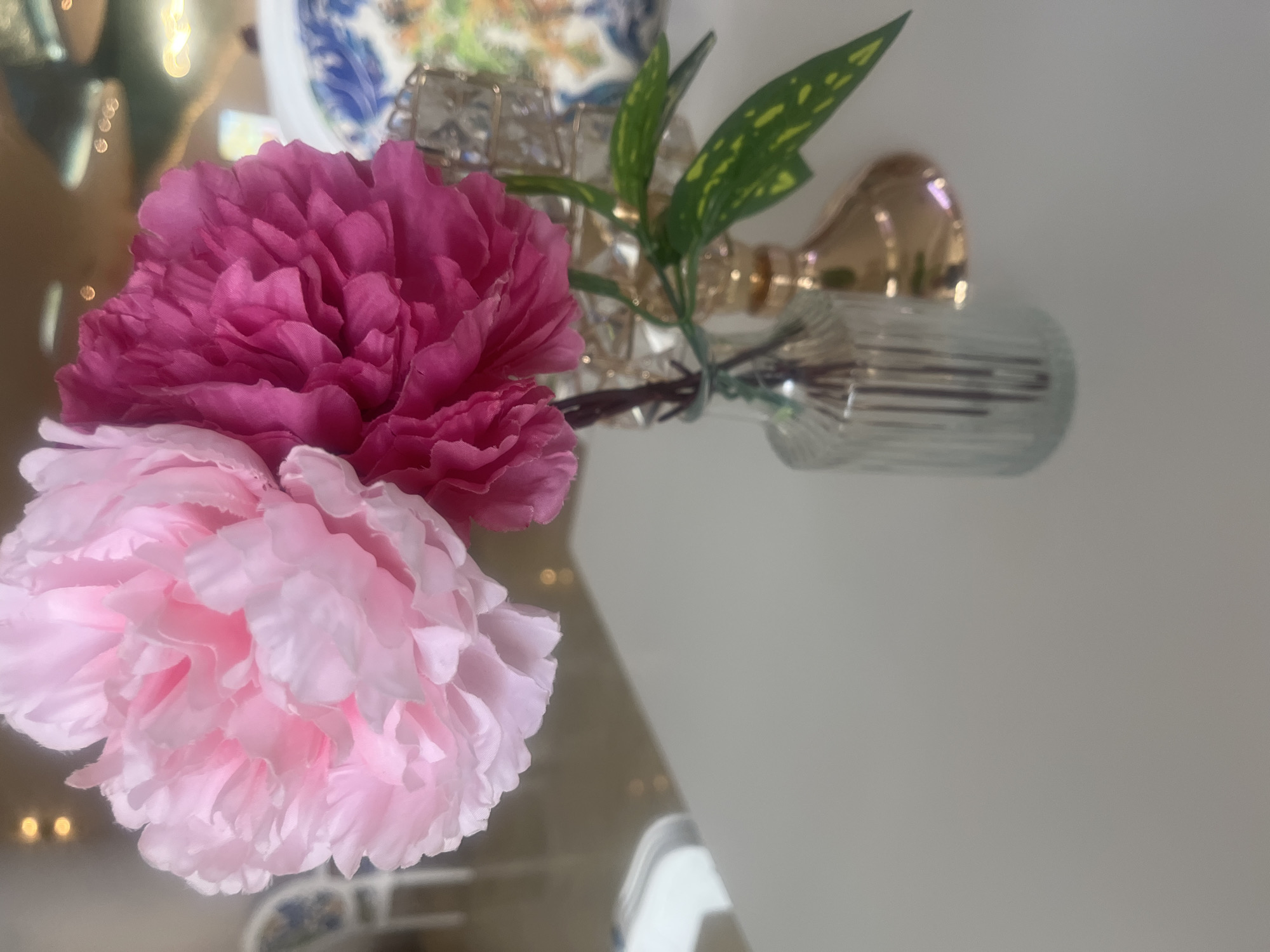 Two artificial peonies, one light pink and one dark pink, are arranged in a small, fluted glass vase on a white surface. The flowers have full, layered petals. A few green leaves with yellow speckles are also visible. Behind the vase, a gold-colored decorative object, possibly a candlestick or stand, is present. In the blurred background, hints of chairs and soft lighting suggest an indoor setting, possibly a room in a building. The scene is lit with soft, diffused light.