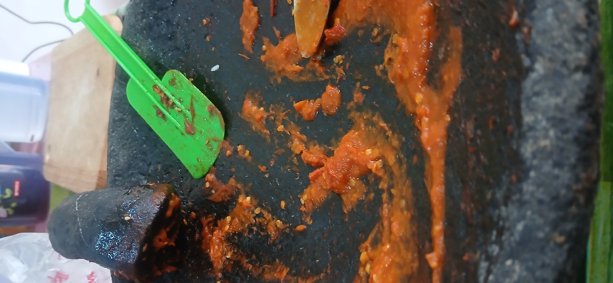 A close-up, vertical, eye-level shot captures a stone mortar and pestle on a dark, rough surface, heavily splattered with a vibrant orange-red chili paste. A bright green spatula rests on the mortar, also smeared with the paste. The background is blurred, showing hints of kitchen items like a wooden cutting board and containers, suggesting an indoor setting. The chili paste is unevenly distributed, with some areas thick and chunky, and others spread thinly, revealing small chili seeds. The lighting is soft, highlighting the textures of the paste and the stone. The scene appears to be located in West Jakarta, Indonesia, given the prevalence of chili paste as a common ingredient in Indonesian cuisine.