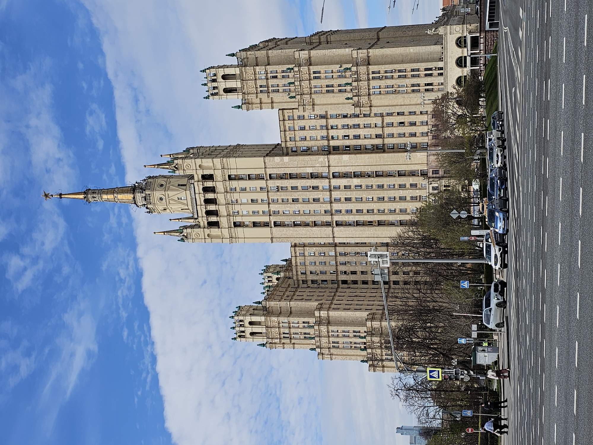 The image depicts the Kotelnicheskaya Embankment Building, a Stalinist skyscraper in Moscow, Russia, viewed from across the street. The building is tall with several tiers and a central spire. The sky above is blue with scattered white clouds. A wide asphalt road is visible in the foreground with white lane markings, several parked cars, and pedestrian crosswalks with signage. Some trees line the street, partly obscuring the lower levels of the building. People are visible crossing the street. The overall scene suggests a clear, sunny day in an urban environment.