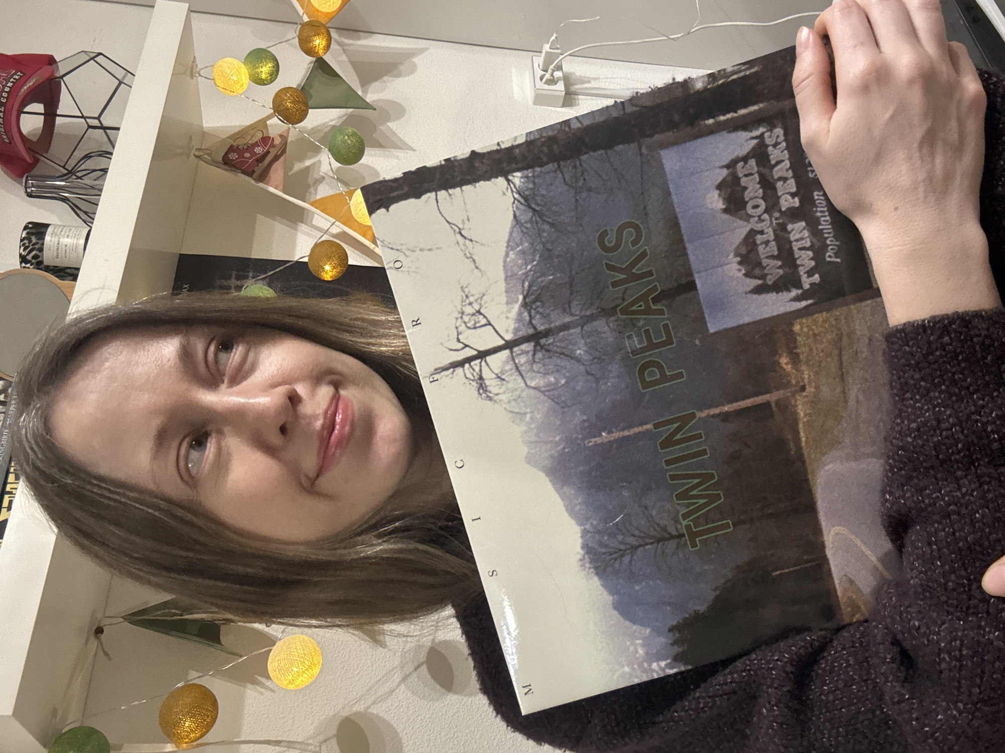 A woman is holding a "Twin Peaks" record album in front of her, with the album cover prominently displayed. The album cover features an image of a forest landscape and the words "WELCOME TO TWIN PEAKS Population." The woman is smiling slightly and looking at the camera. In the background, there is a white shelf with various objects on it, including a red baseball cap, a wire geometric object, a bottle, and string lights with colorful spheres. The setting appears to be indoors. There is no indication that this image was taken in the city of Kudrovo, Russia.